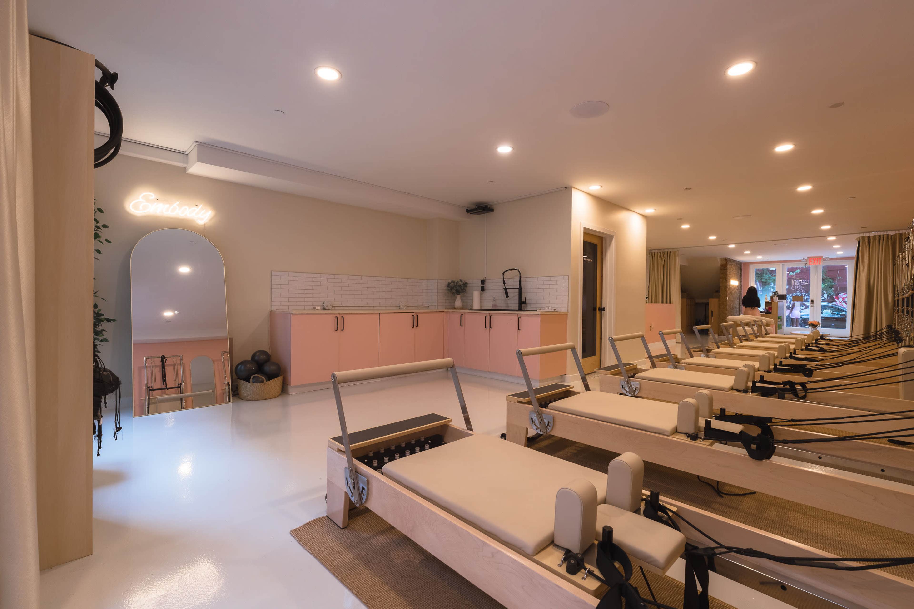 The image depicts a modern studio designed for pilates, featuring multiple reformer machines arranged neatly in a spacious, light-colored room with soft pink accents.