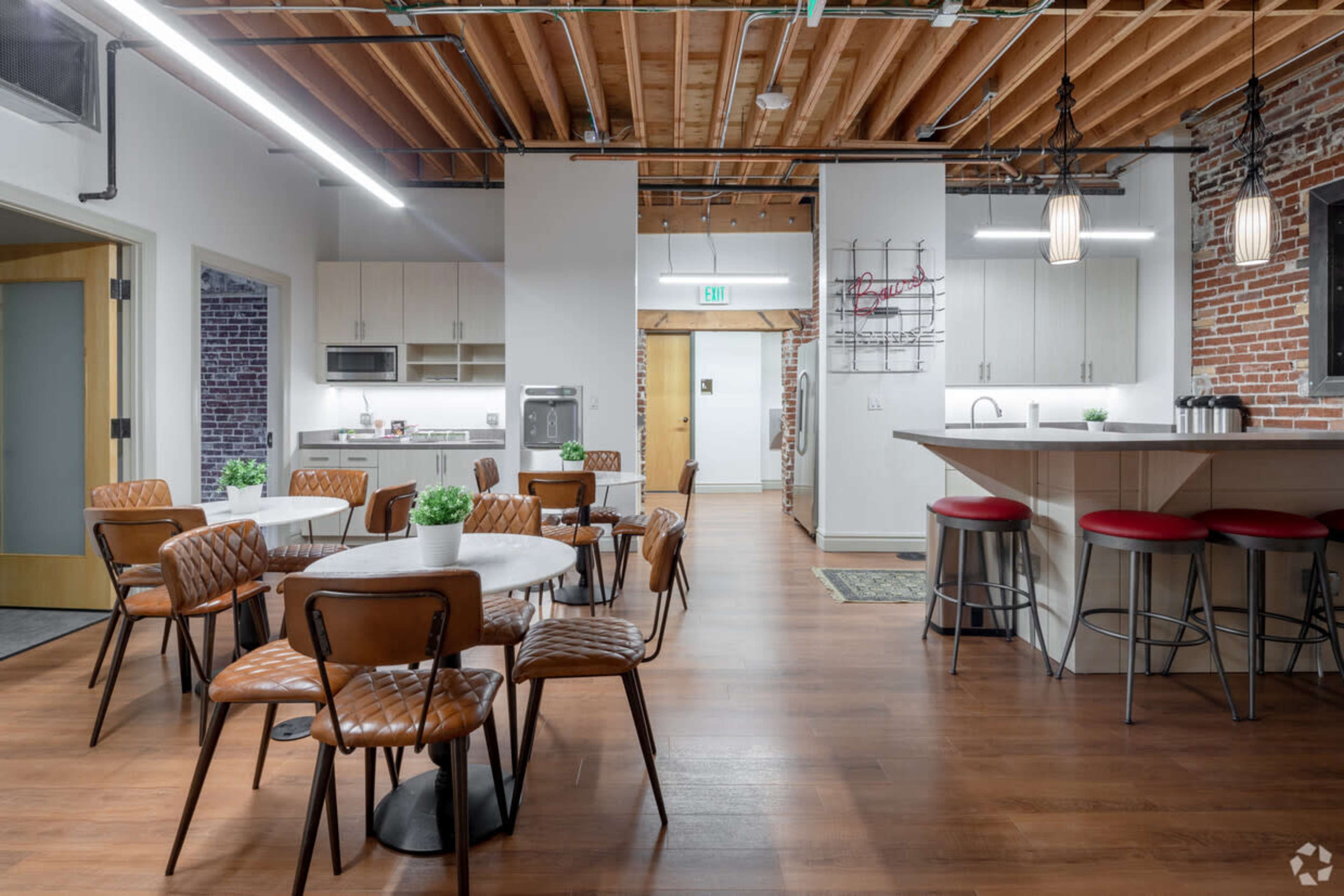 The image shows a modern, open kitchen and dining area with wooden floors, stainless steel appliances, and a mix of round and rectangular tables surrounded by chairs.