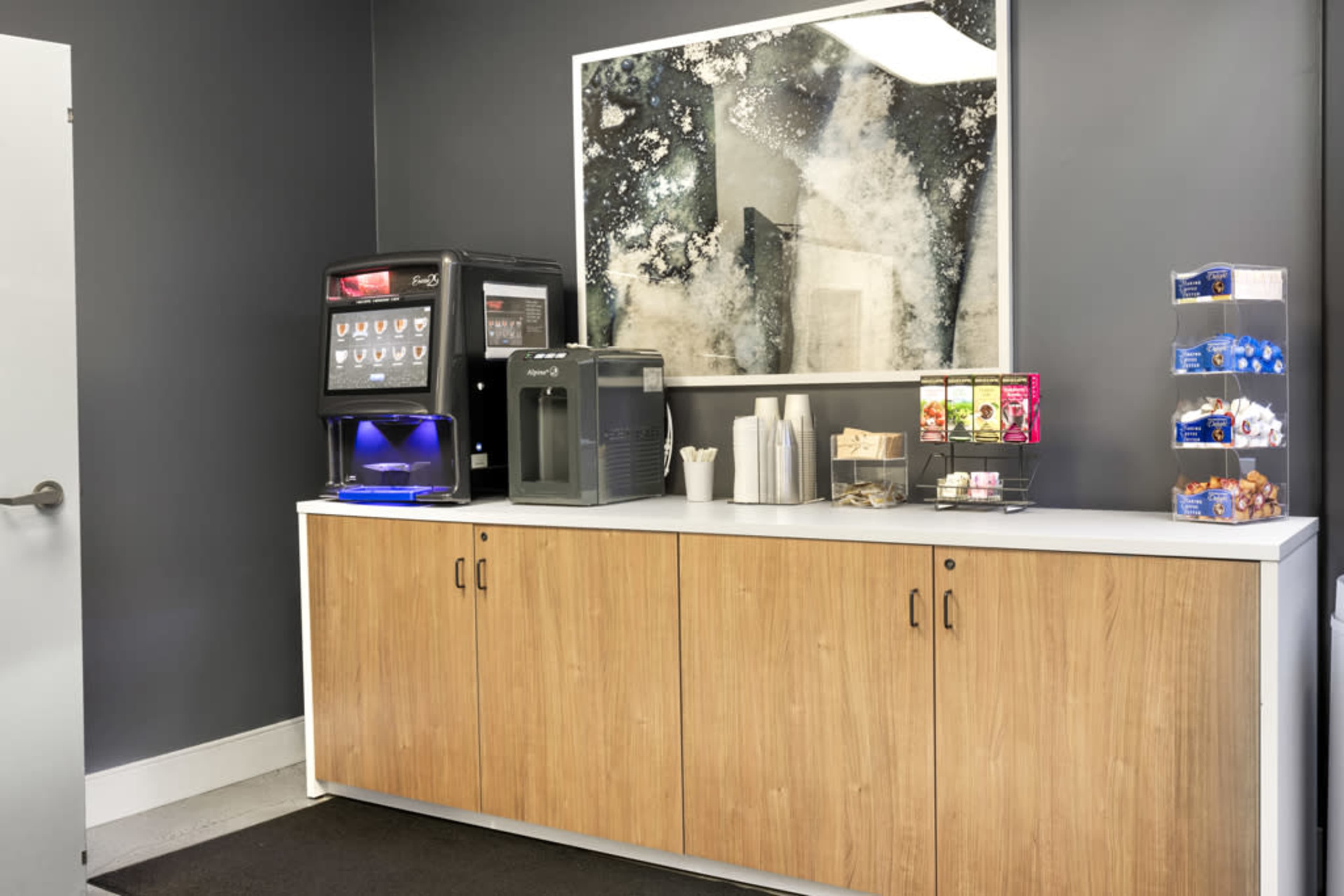 The image shows a snack area with a beverage dispenser, a water cooler, and a selection of snacks arranged on a counter beneath a large abstract wall art piece.