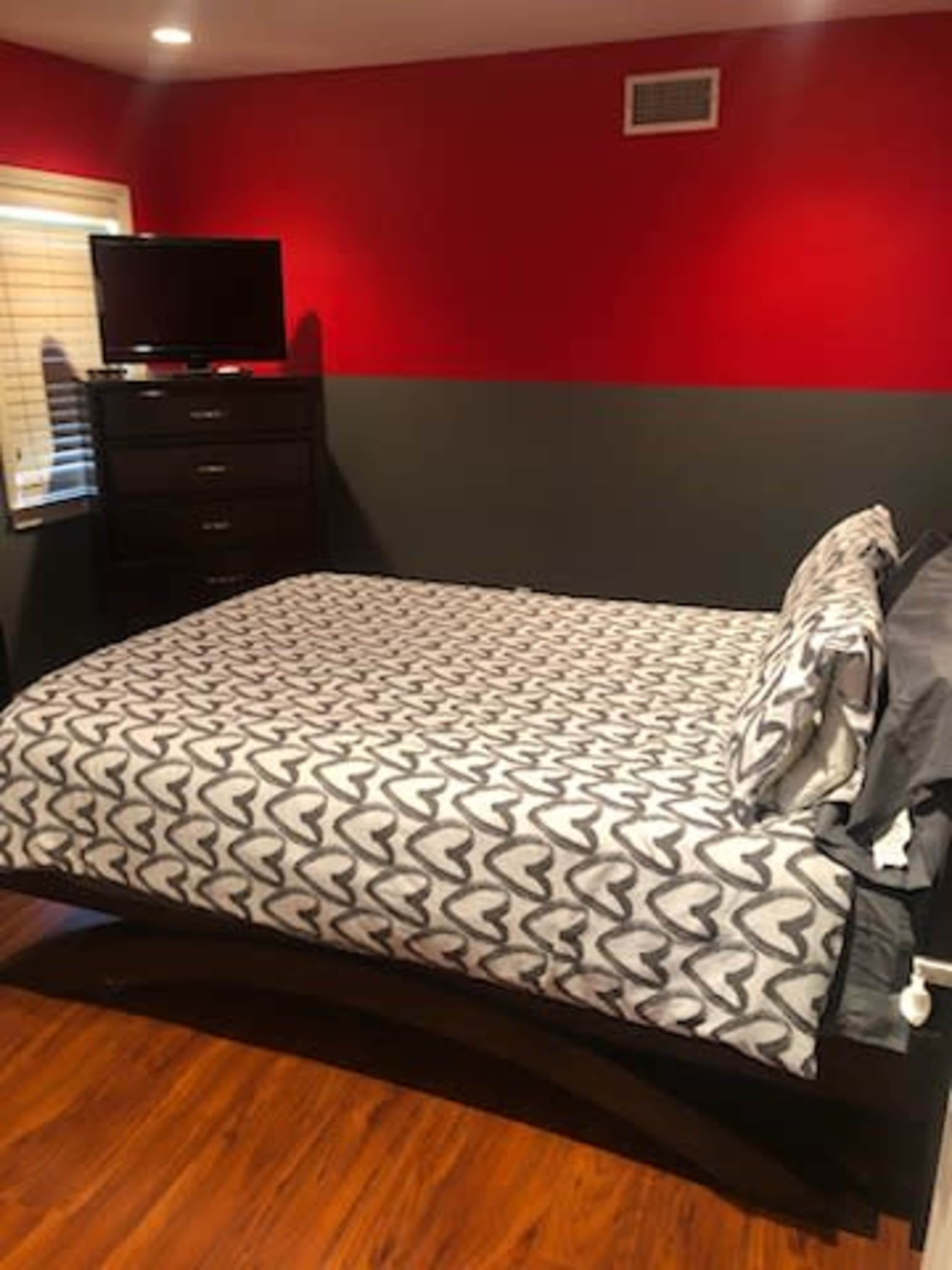 The image shows a bedroom with a red and gray color scheme, featuring a bed with a patterned comforter, a dresser, and a wall-mounted television.