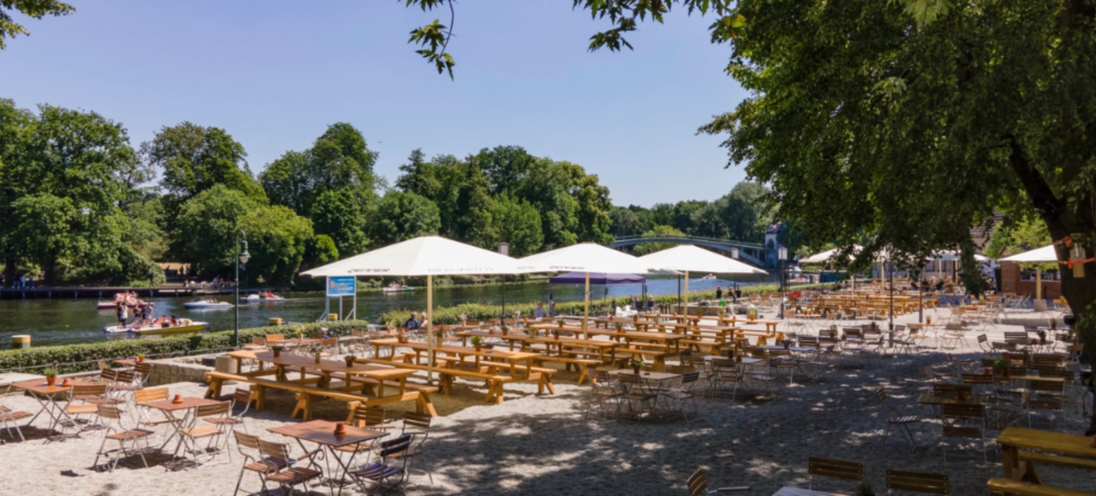 A riverside area features rows of wooden tables and chairs under large umbrellas, with trees lining the bank and boats visible on the water.