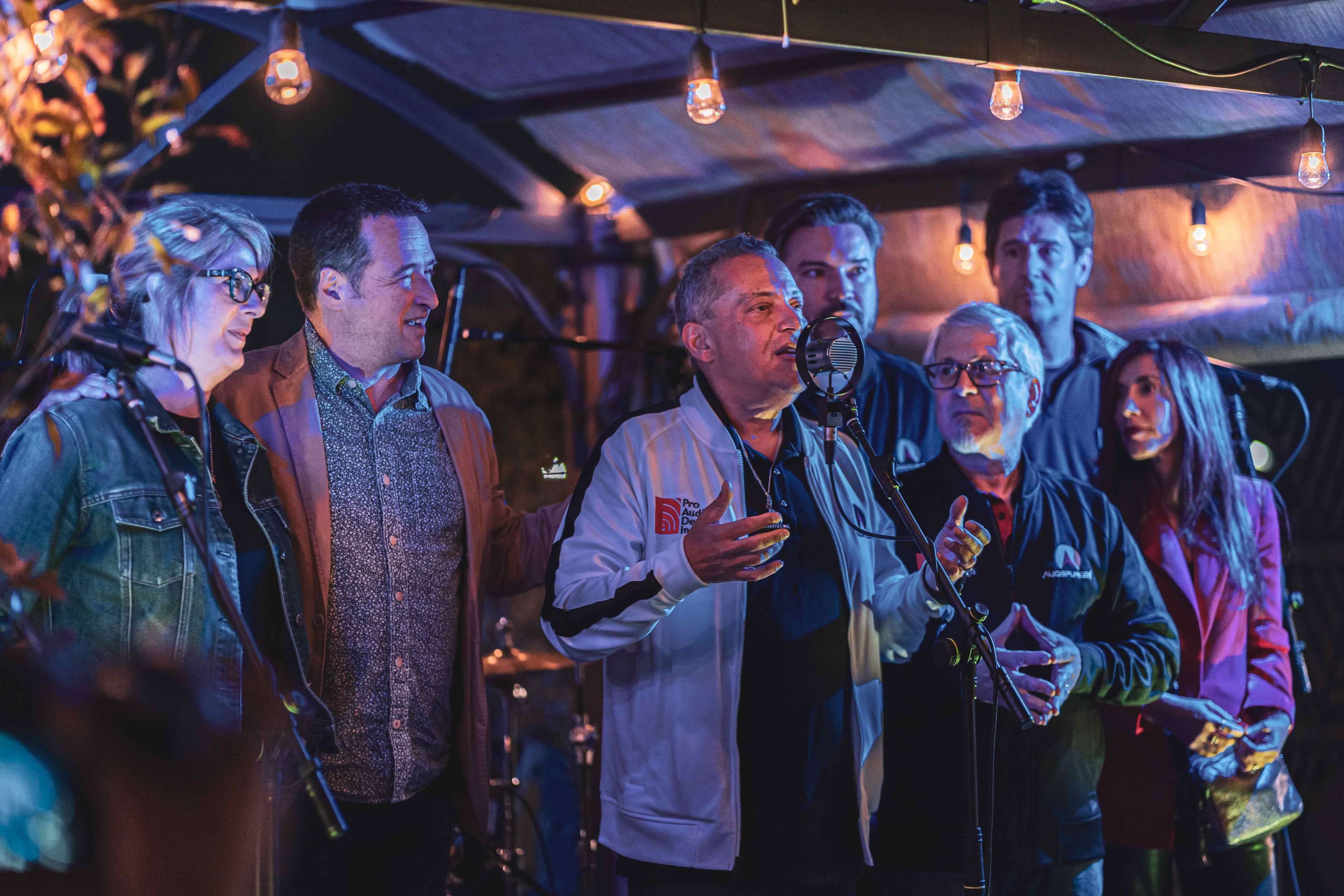A group of people stands under a canopy with string lights, as one man speaks into a microphone while others listen attentively.