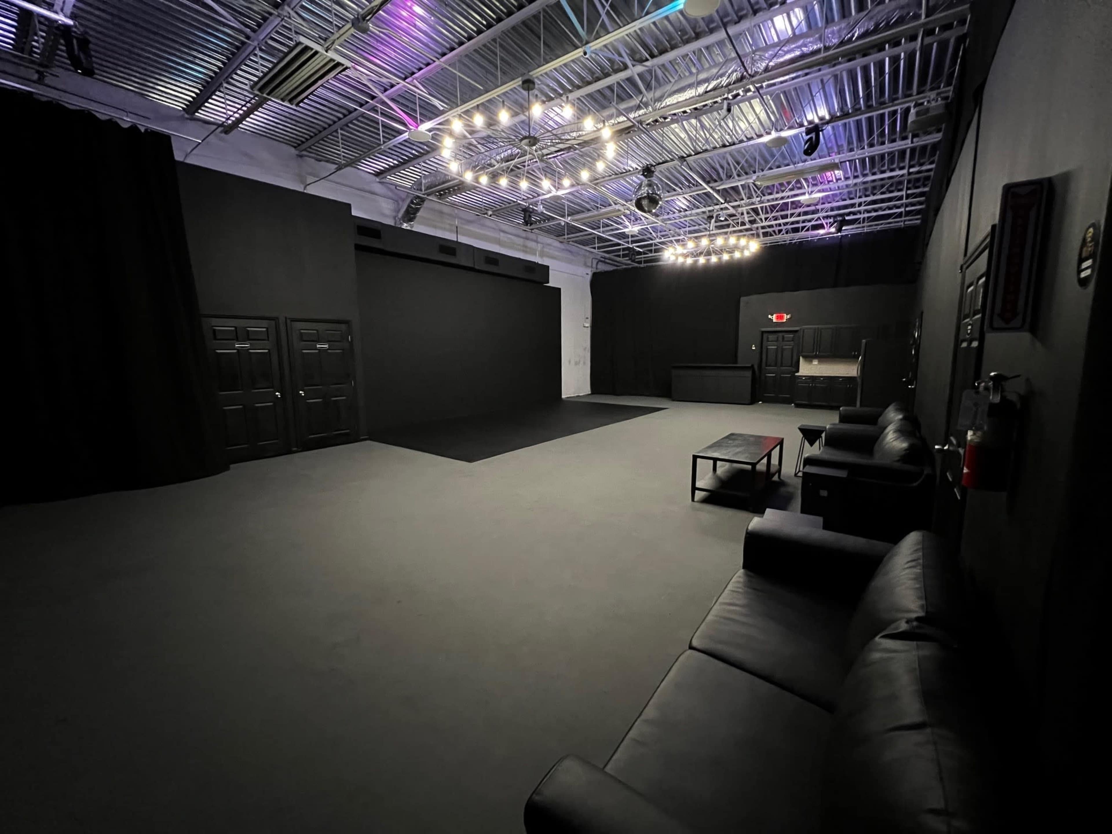 A spacious black-walled studio with a flat floor, several couches along one side, and overhead lights illuminating the area.