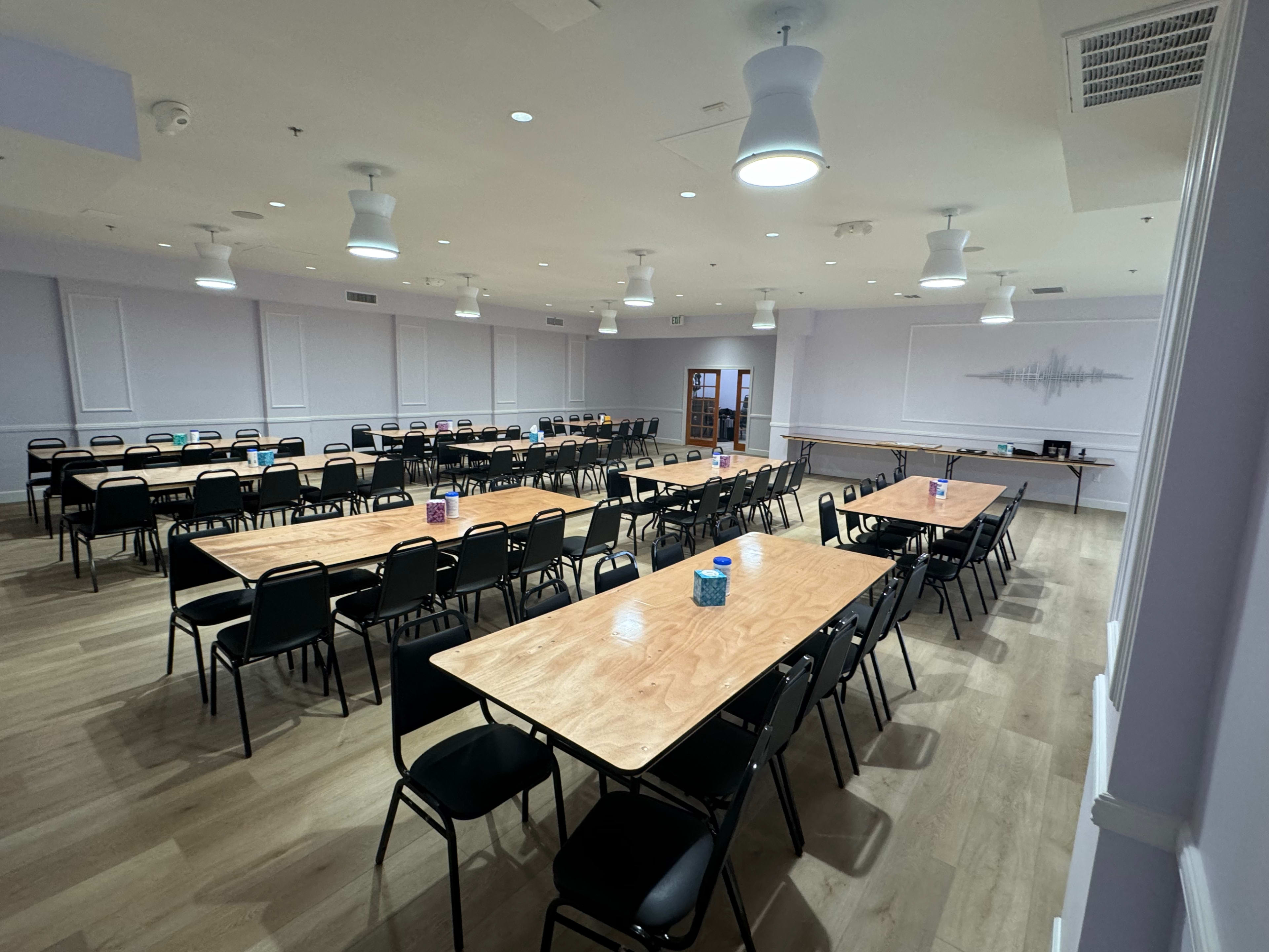 A spacious, well-lit meeting room with multiple rows of wooden tables and black chairs arranged for an event.