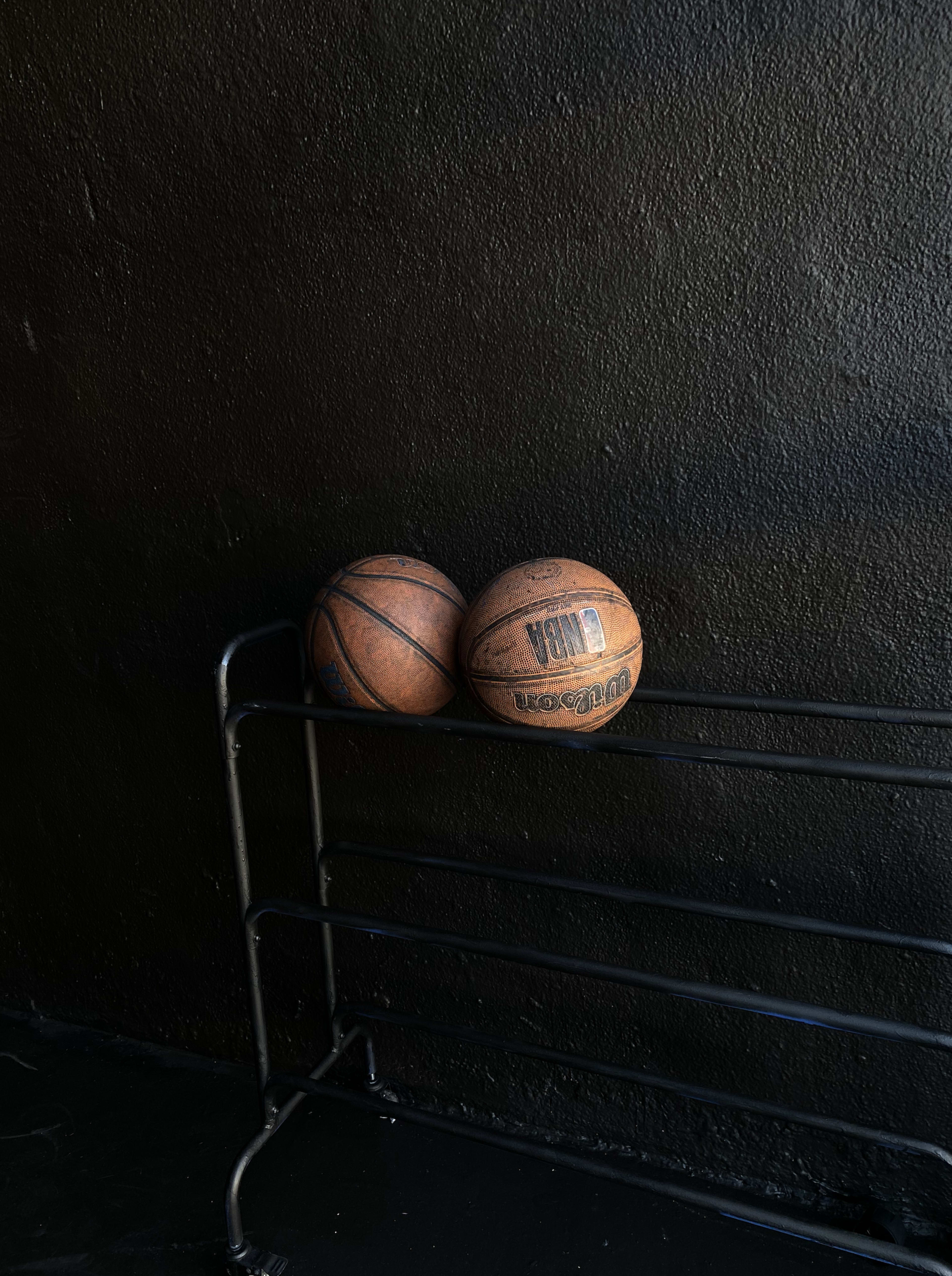 Two worn basketballs rest on a black metal rack against a dark wall.