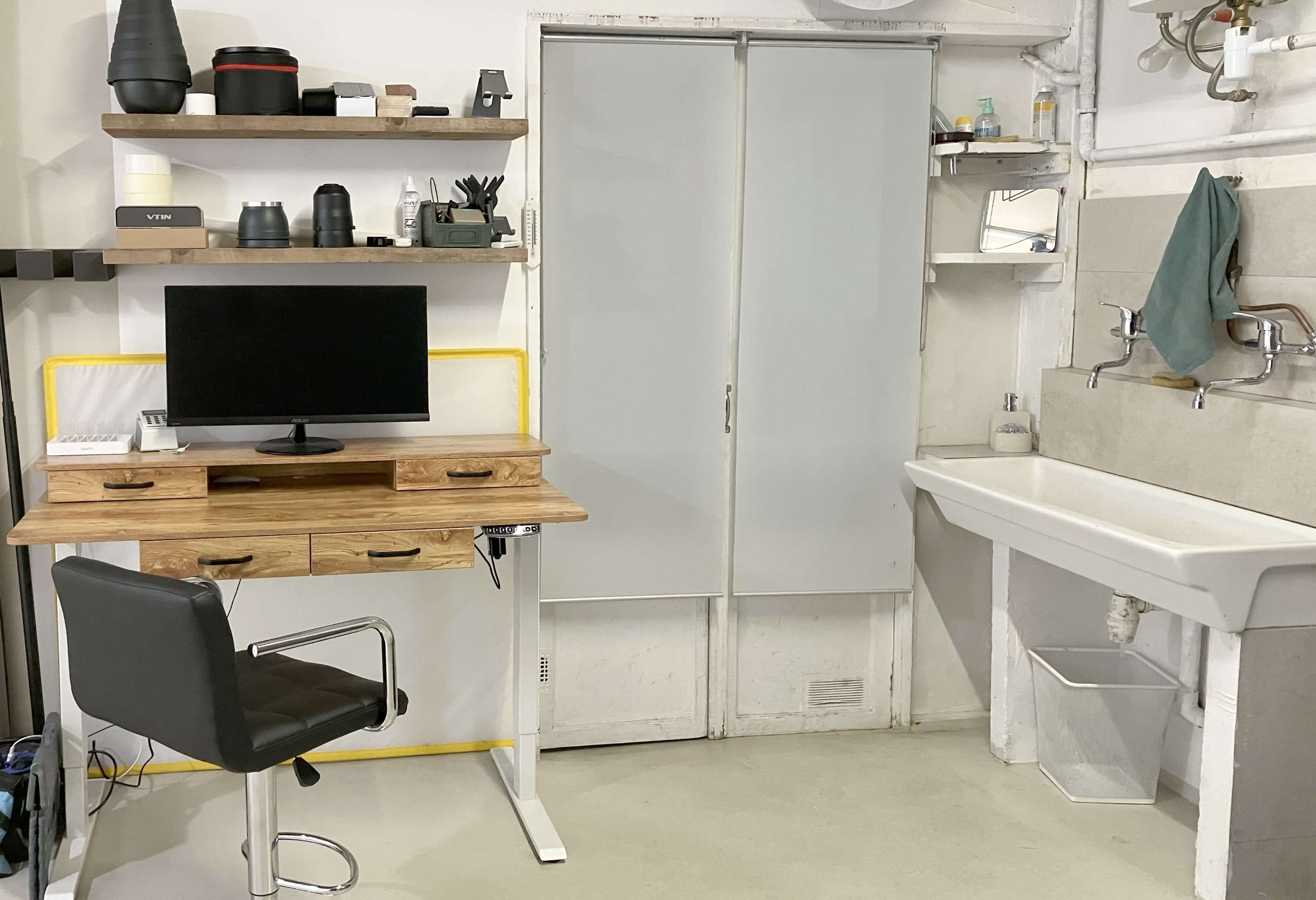 A minimalist workspace with a wooden desk, a monitor, and a black chair, alongside a sink and storage shelves against a wall.