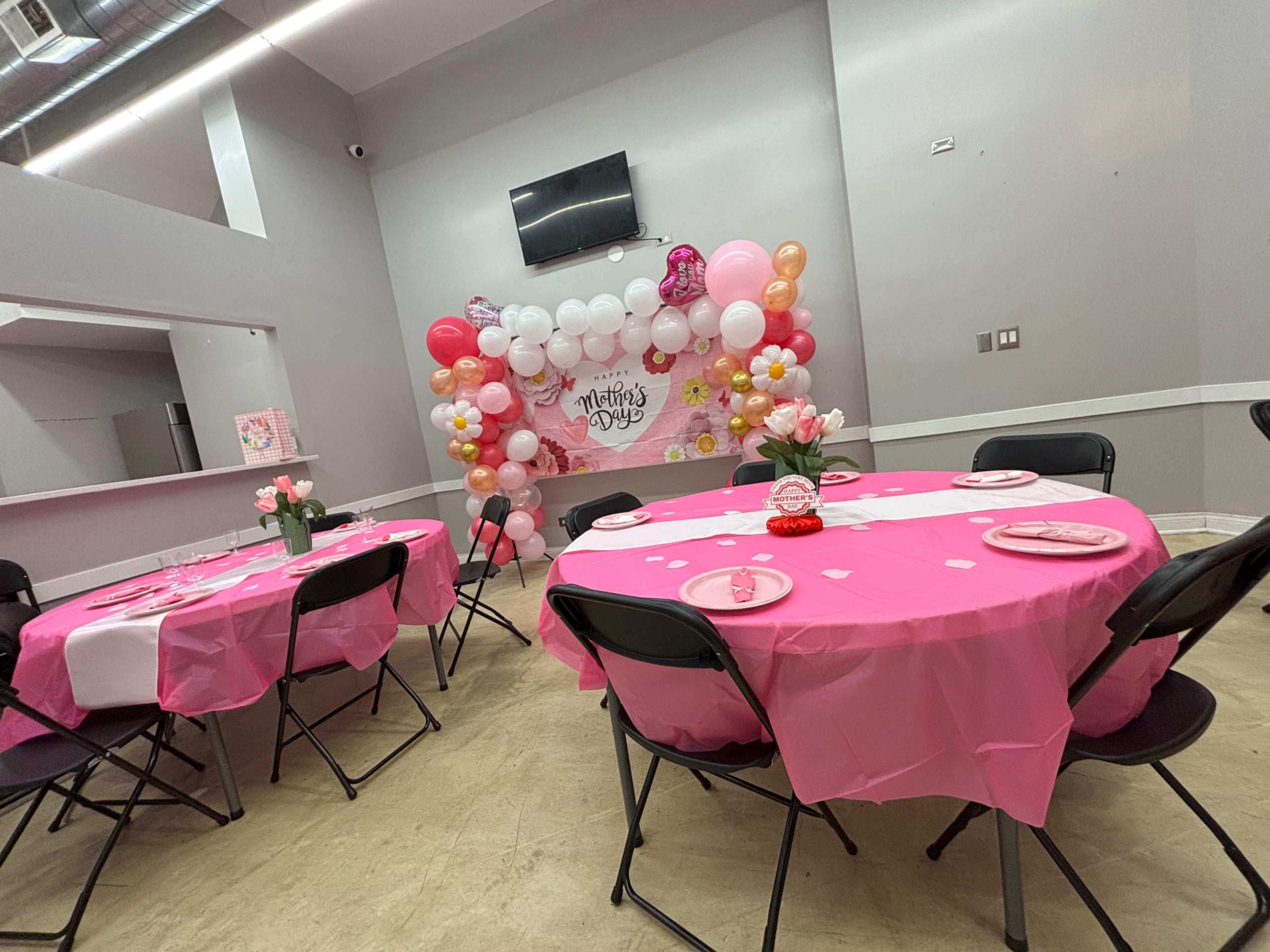 The image shows a decorated room set up for a Mother's Day celebration, featuring pink tablecloths, black chairs, and a balloon backdrop.