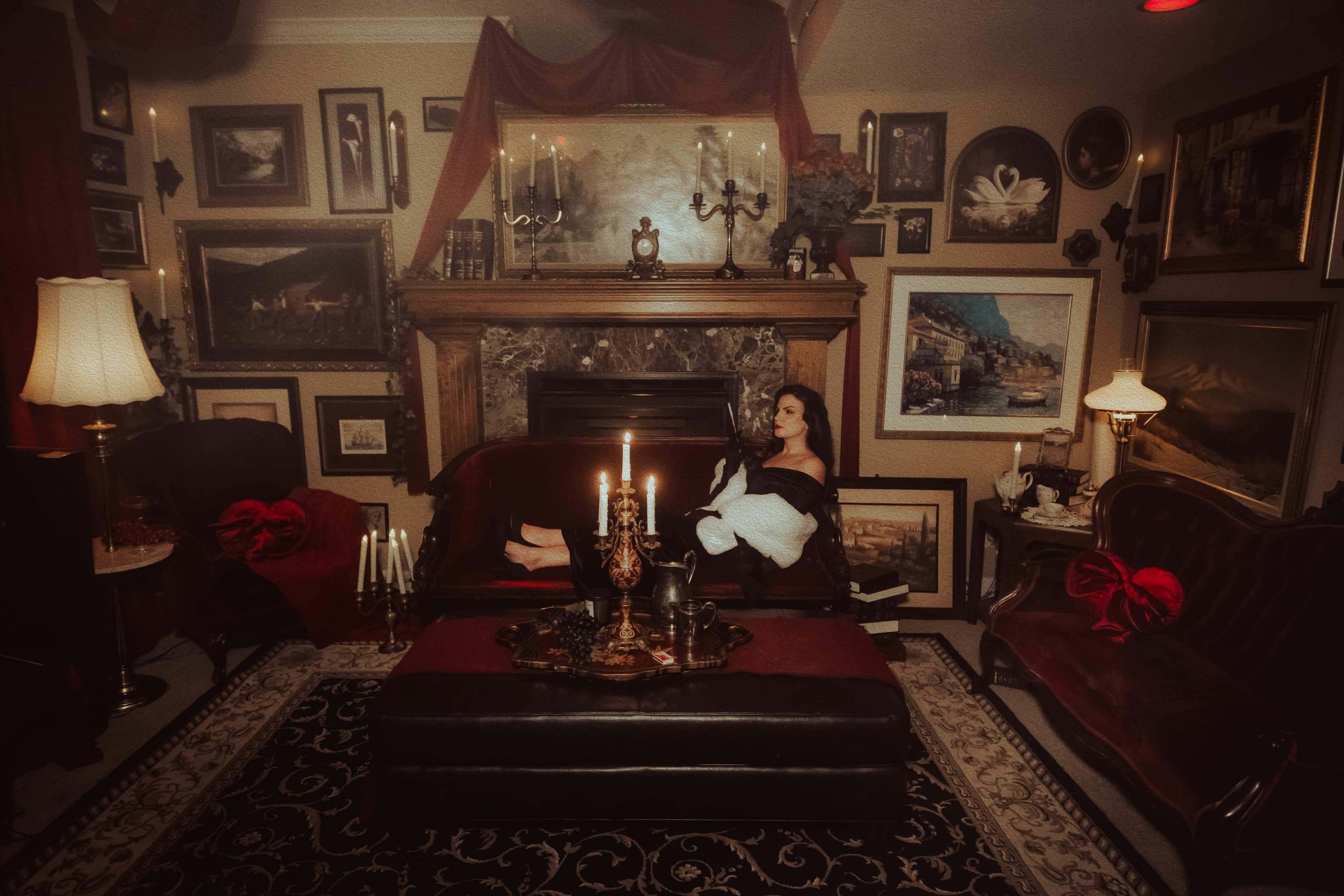 A woman sits on a couch in a richly decorated room filled with vintage artwork and ornate furnishings.