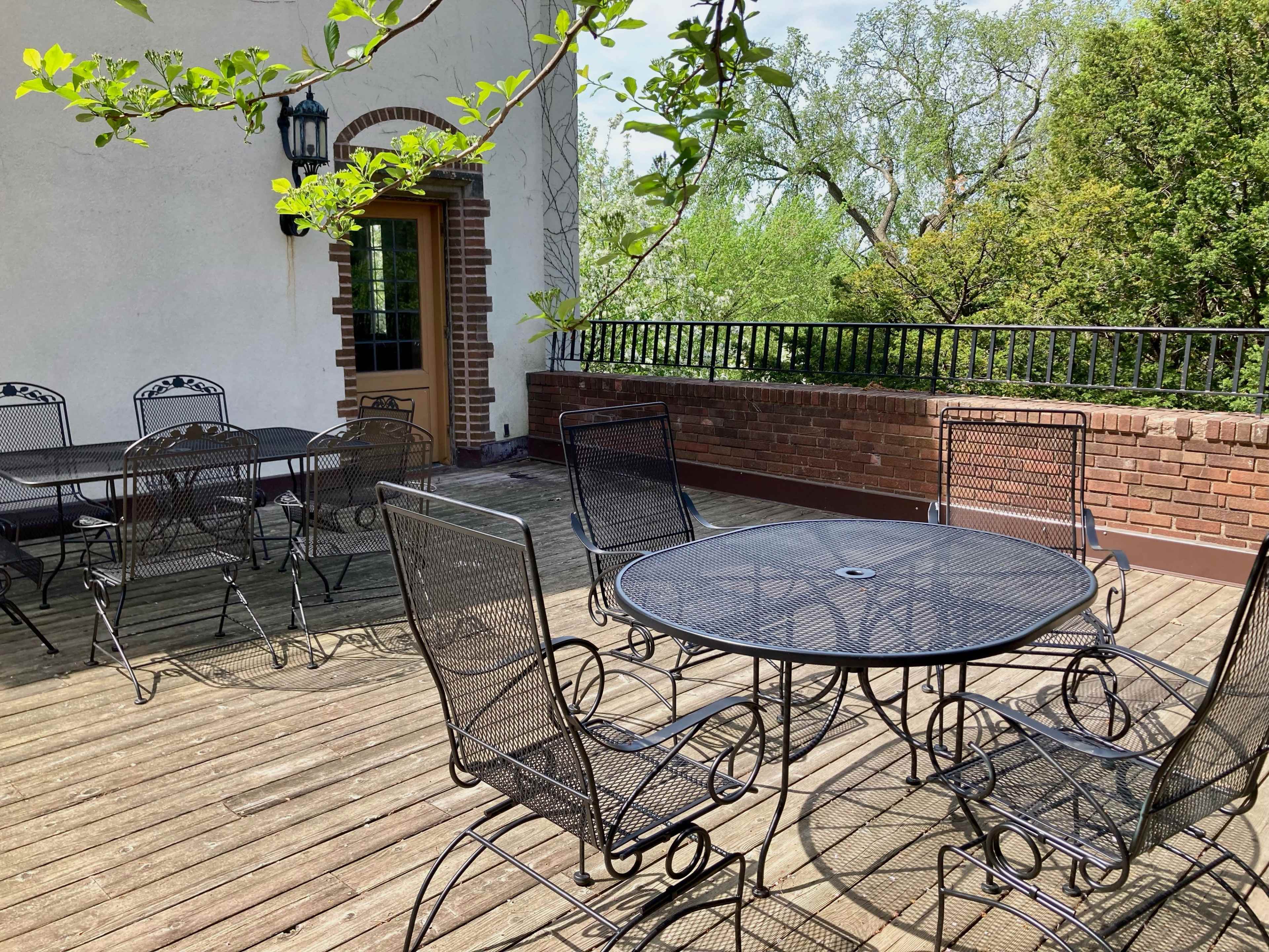 A spacious outdoor patio featuring several black wrought iron tables and chairs arranged on a wooden deck surrounded by greenery.