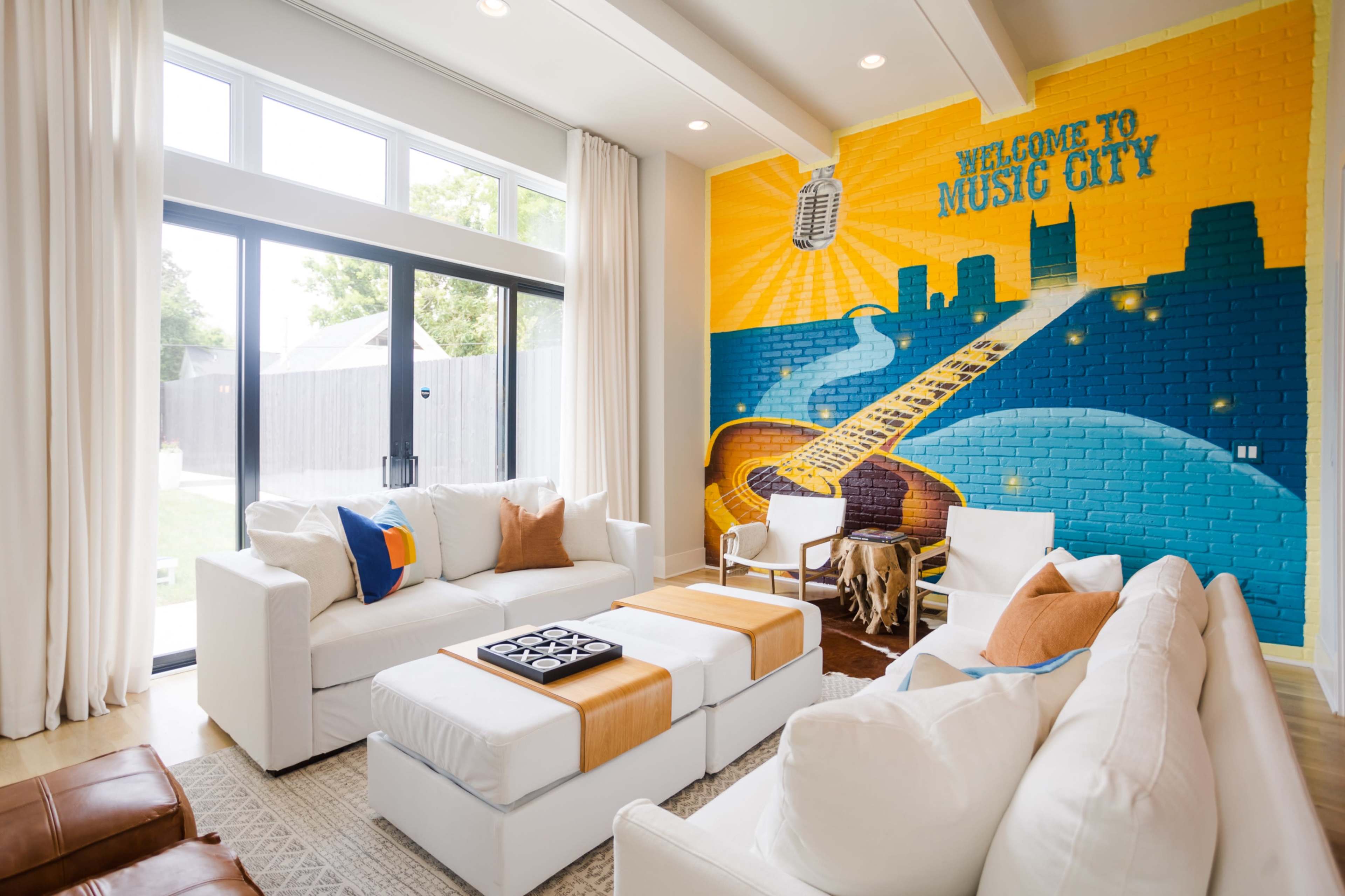 The image shows a bright living room with white furniture, large windows, and a vibrant mural that features a guitar and the words "Welcome to Music City."