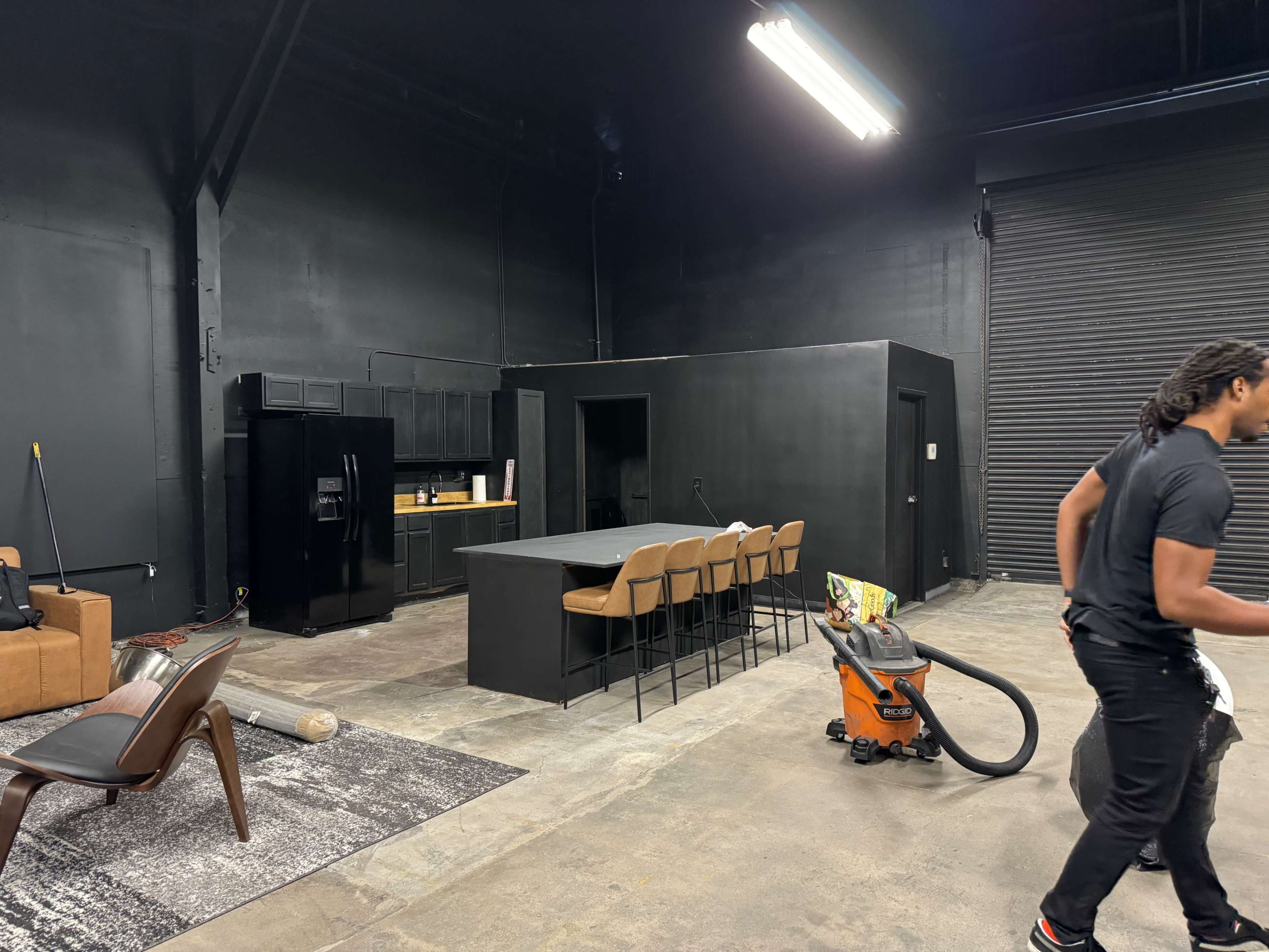 The image shows a spacious, industrial-style room with black walls featuring a kitchen area with bar stools, a dining table, and a person using a vacuum cleaner.