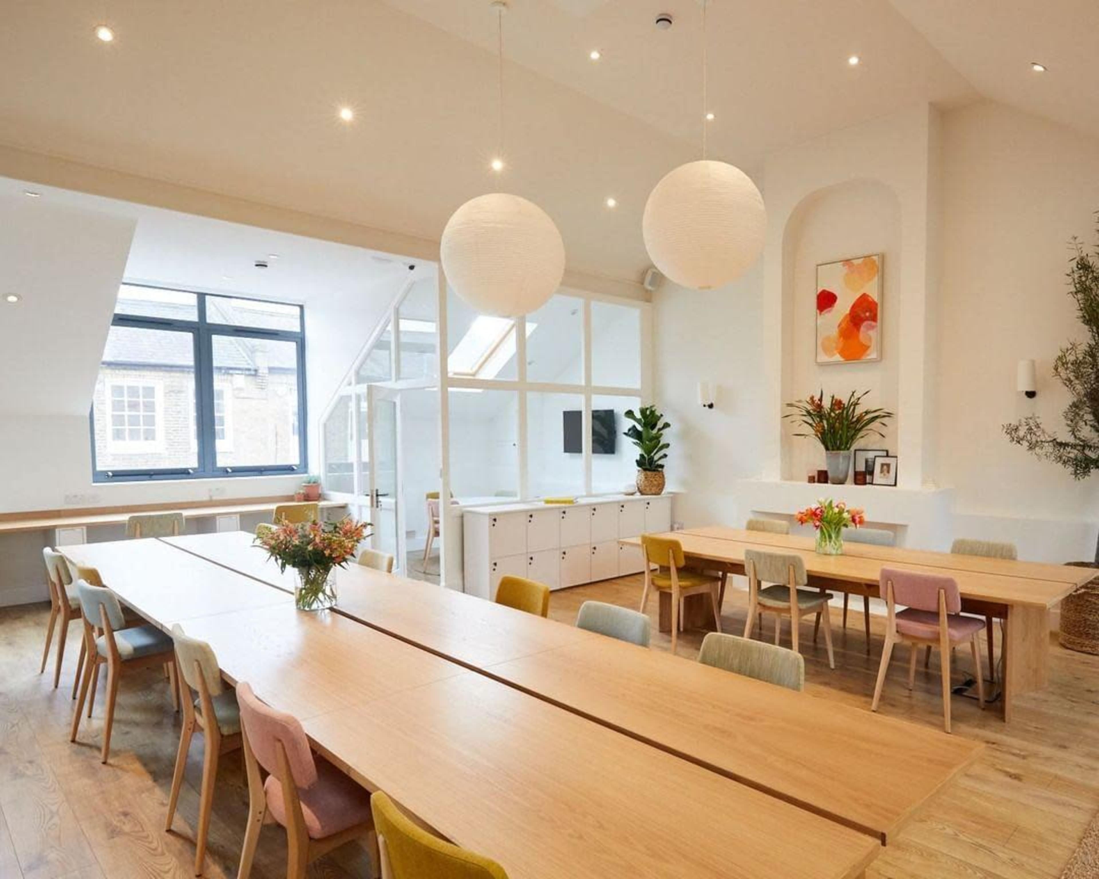 A spacious and well-lit meeting room features a long wooden table with colorful chairs, large windows, and minimalist decor.