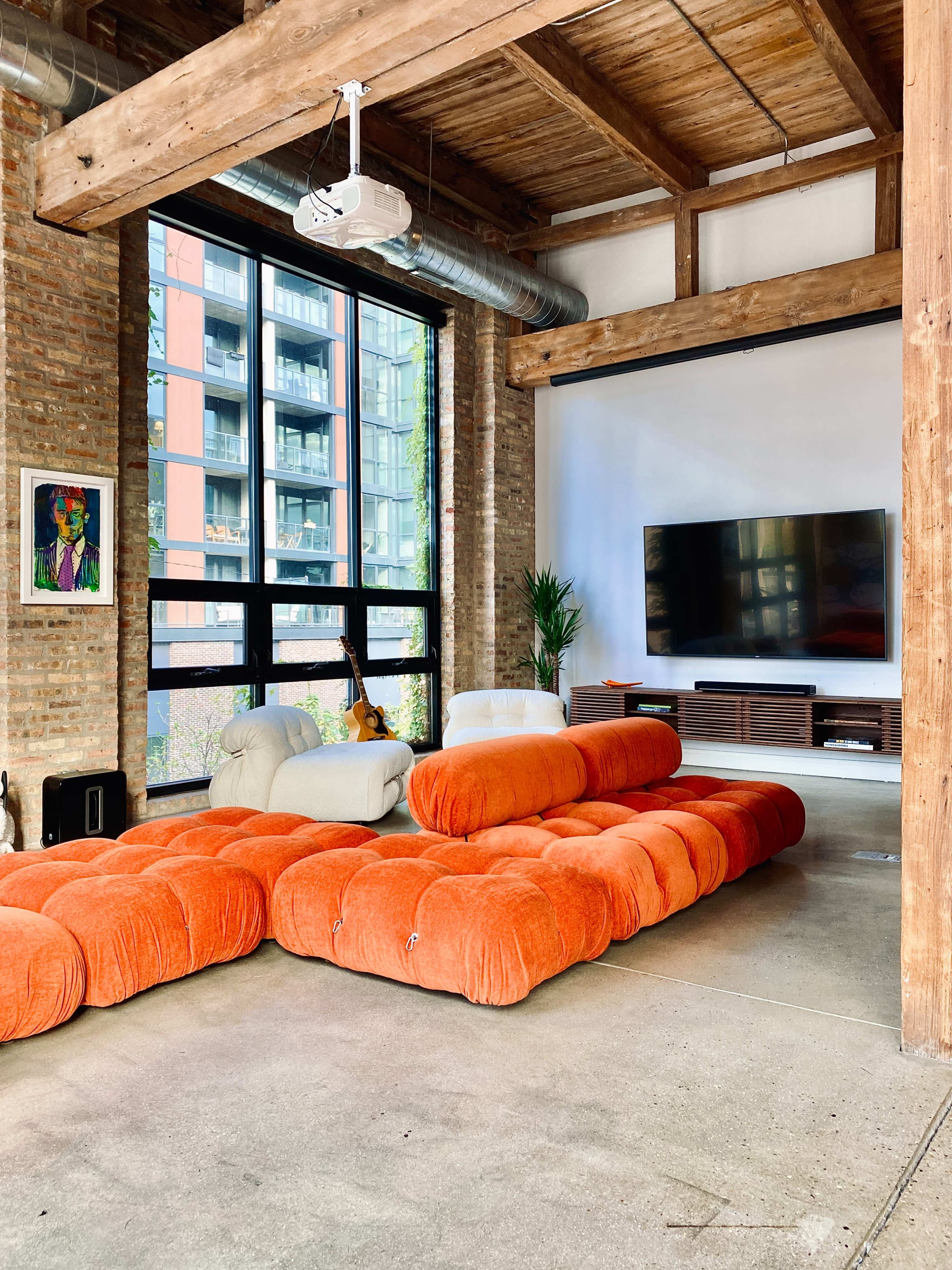 A modern living room with an orange sectional sofa, wooden beams, exposed brick walls, a large television mounted on a wall, and a projector on the ceiling.