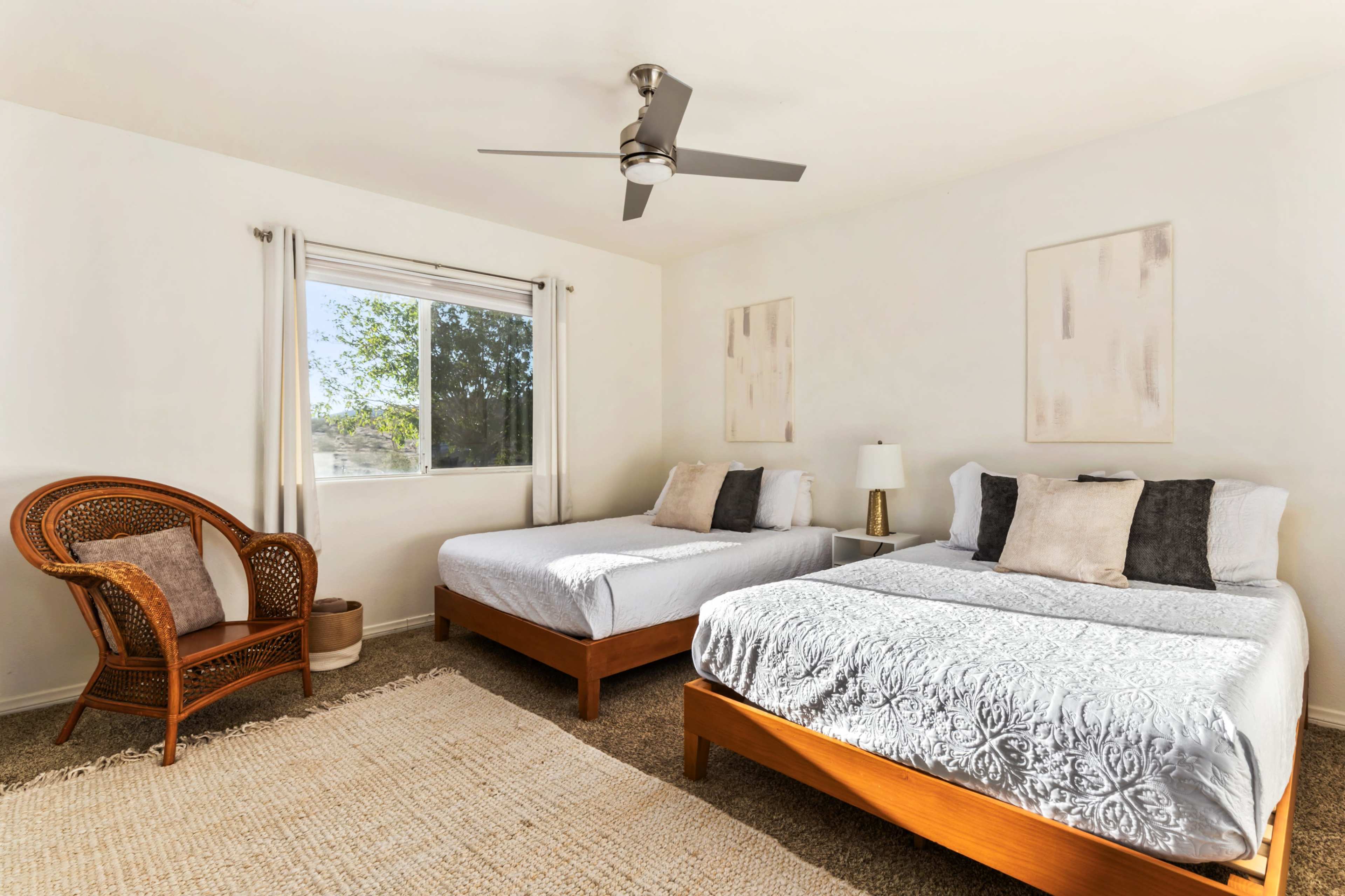 The image shows a bright bedroom with two beds, a wicker chair, a lamp, and a window overlooking greenery.