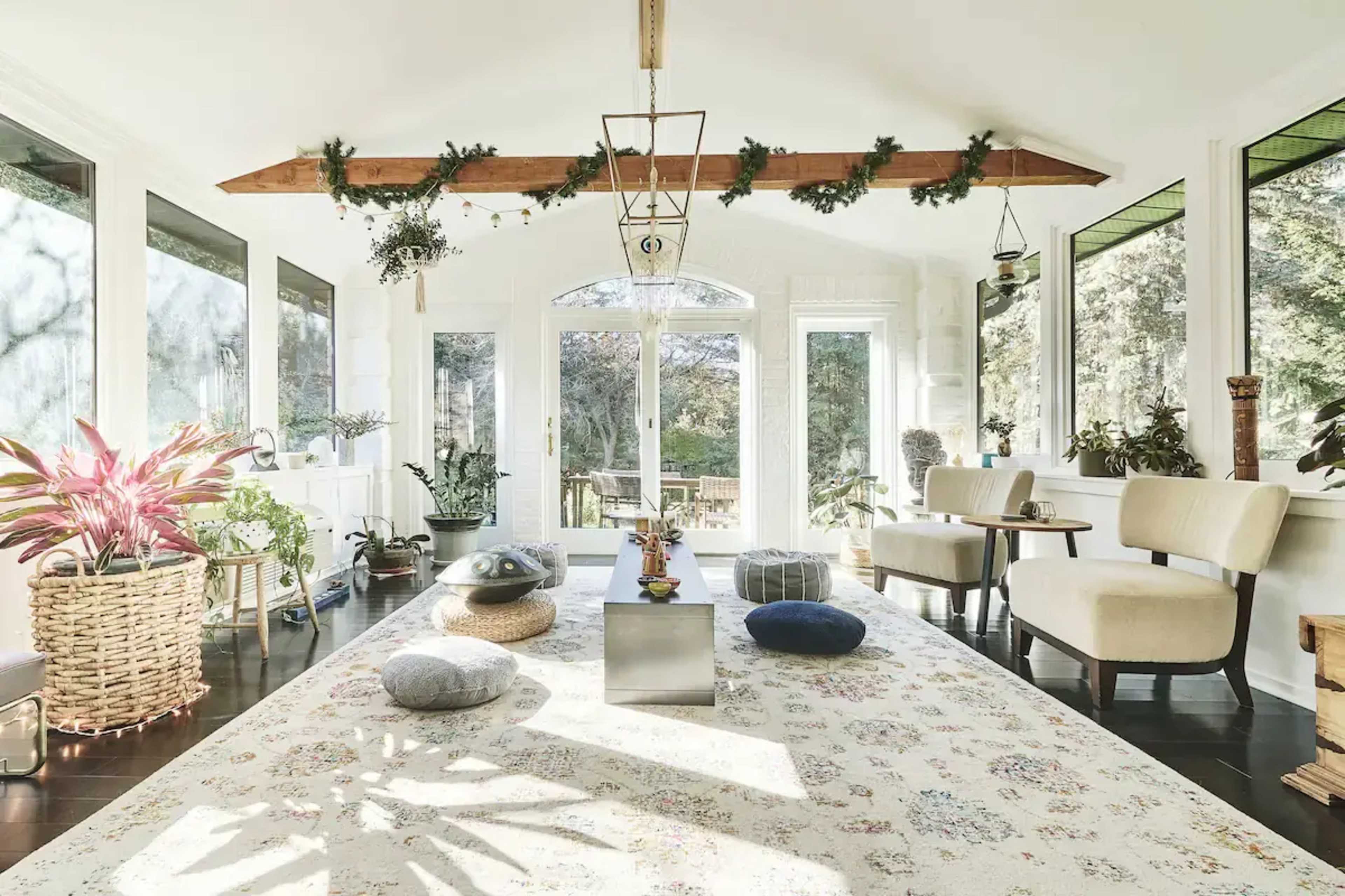 A well-lit sunroom features a patterned rug, various plants, and two chairs arranged around a central table.