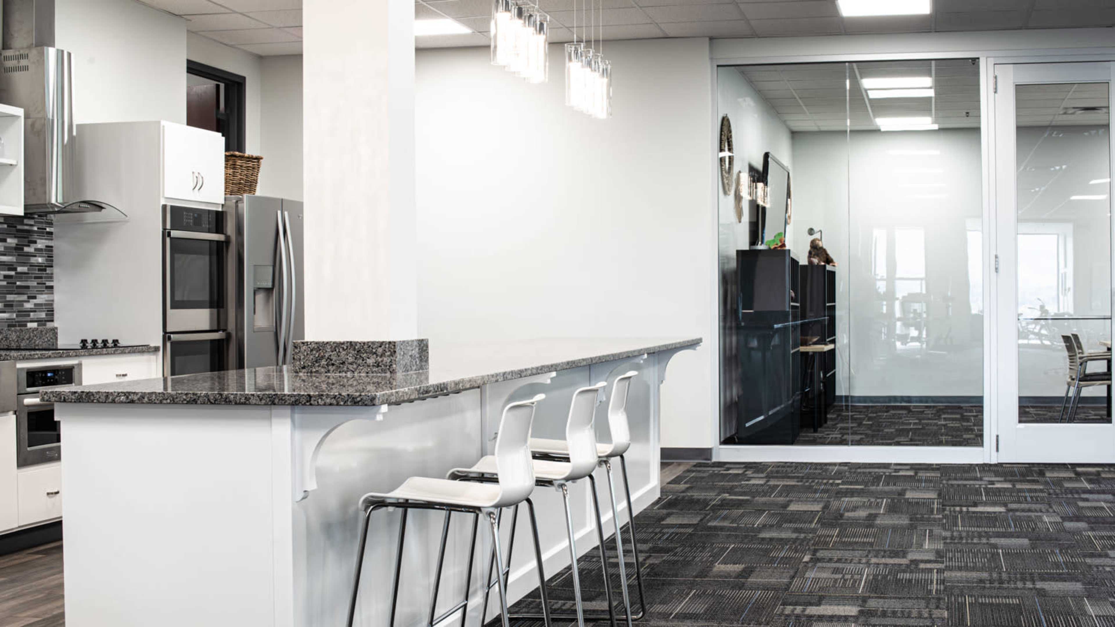 A modern kitchen area with a bar counter, three white stools, and a glass-walled conference room in the background.