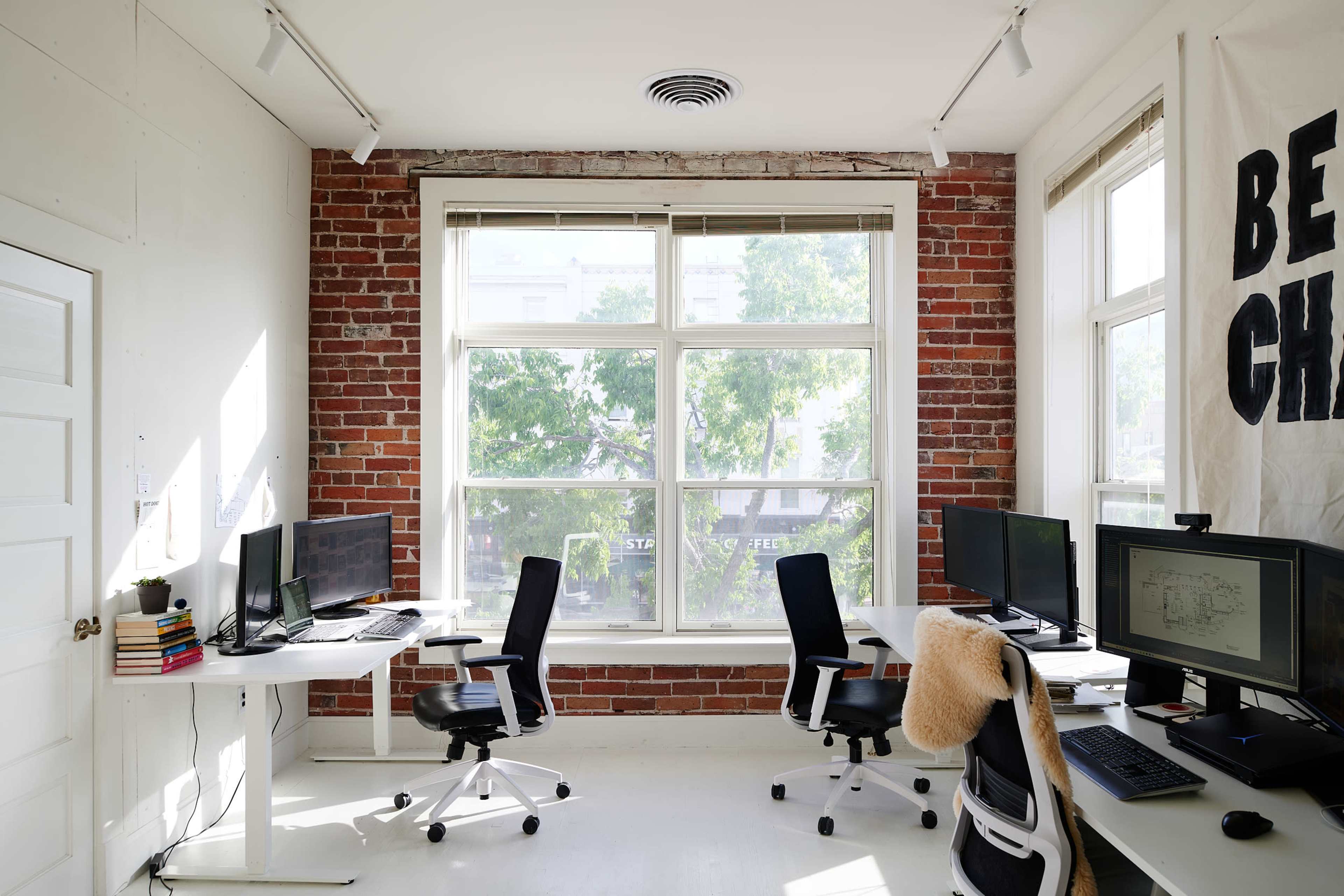 A bright office space with two workstations facing large windows, featuring exposed brick walls and modern furniture.