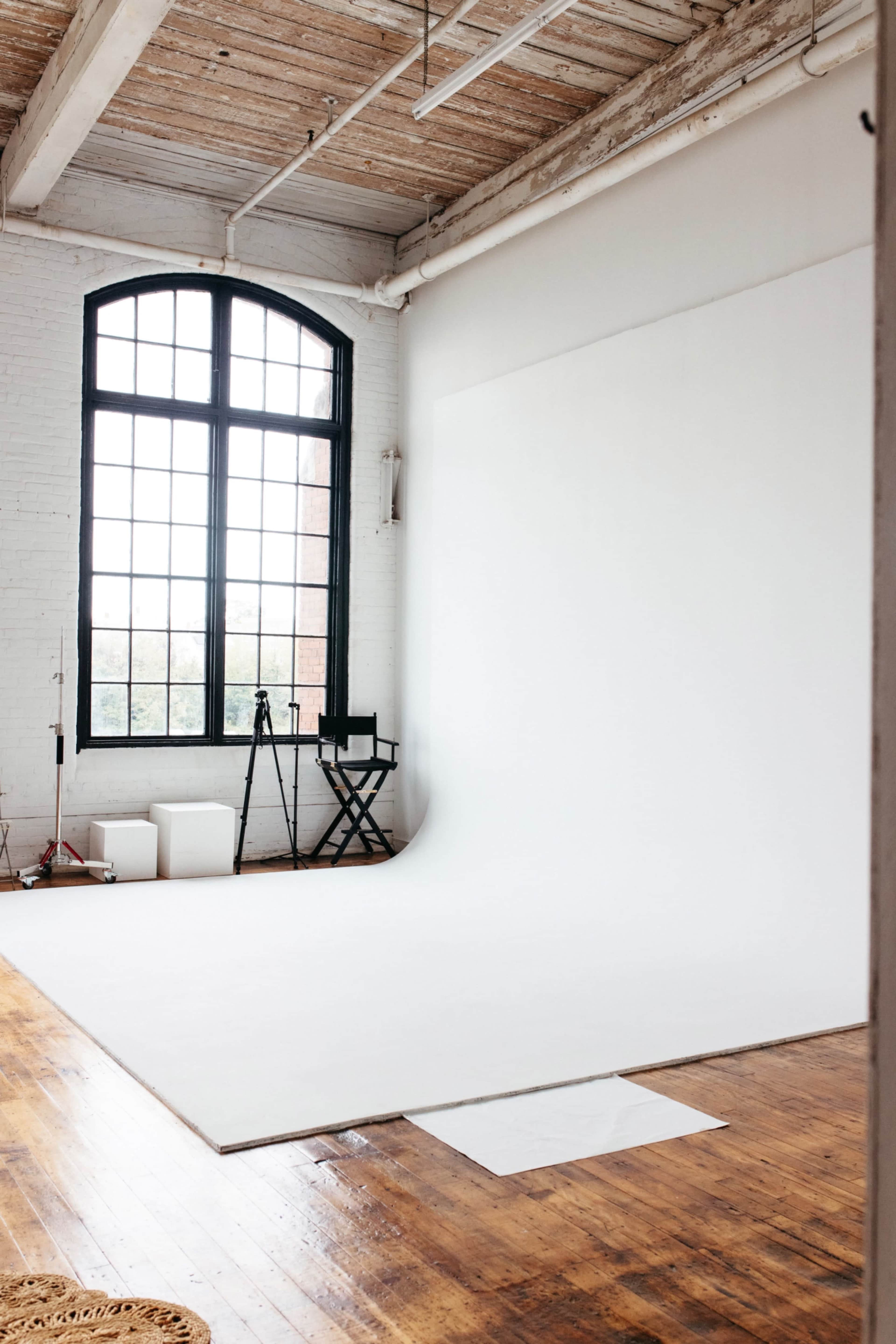 The image shows a spacious, well-lit photography studio with a large window, a white backdrop, and wooden floors.