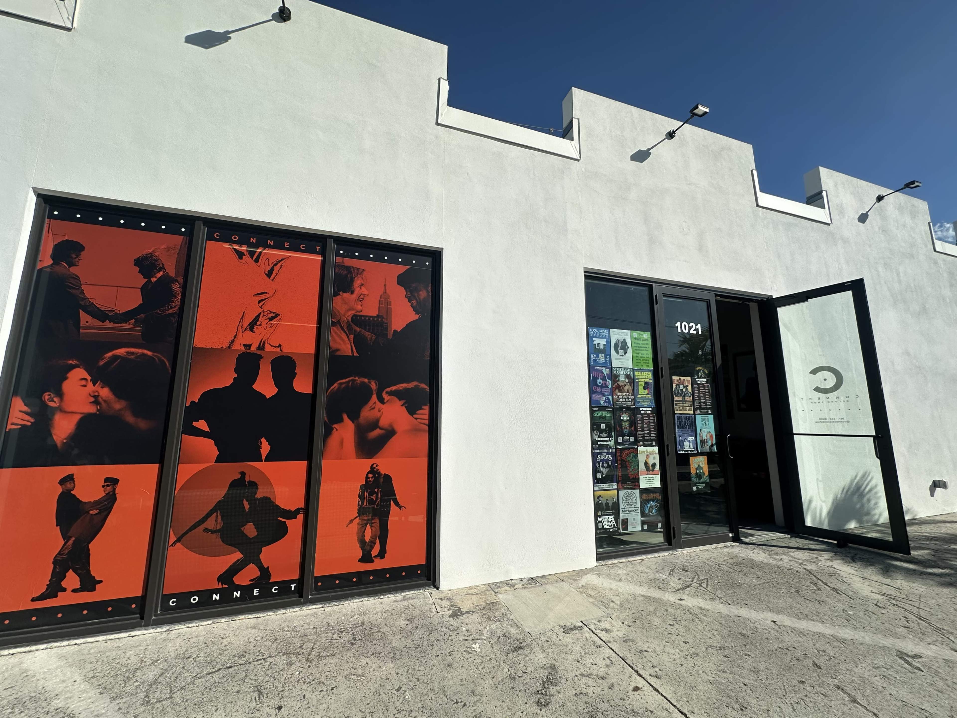 The image shows a white building with large glass doors displaying various posters and an artistic mural in red and black on the side.