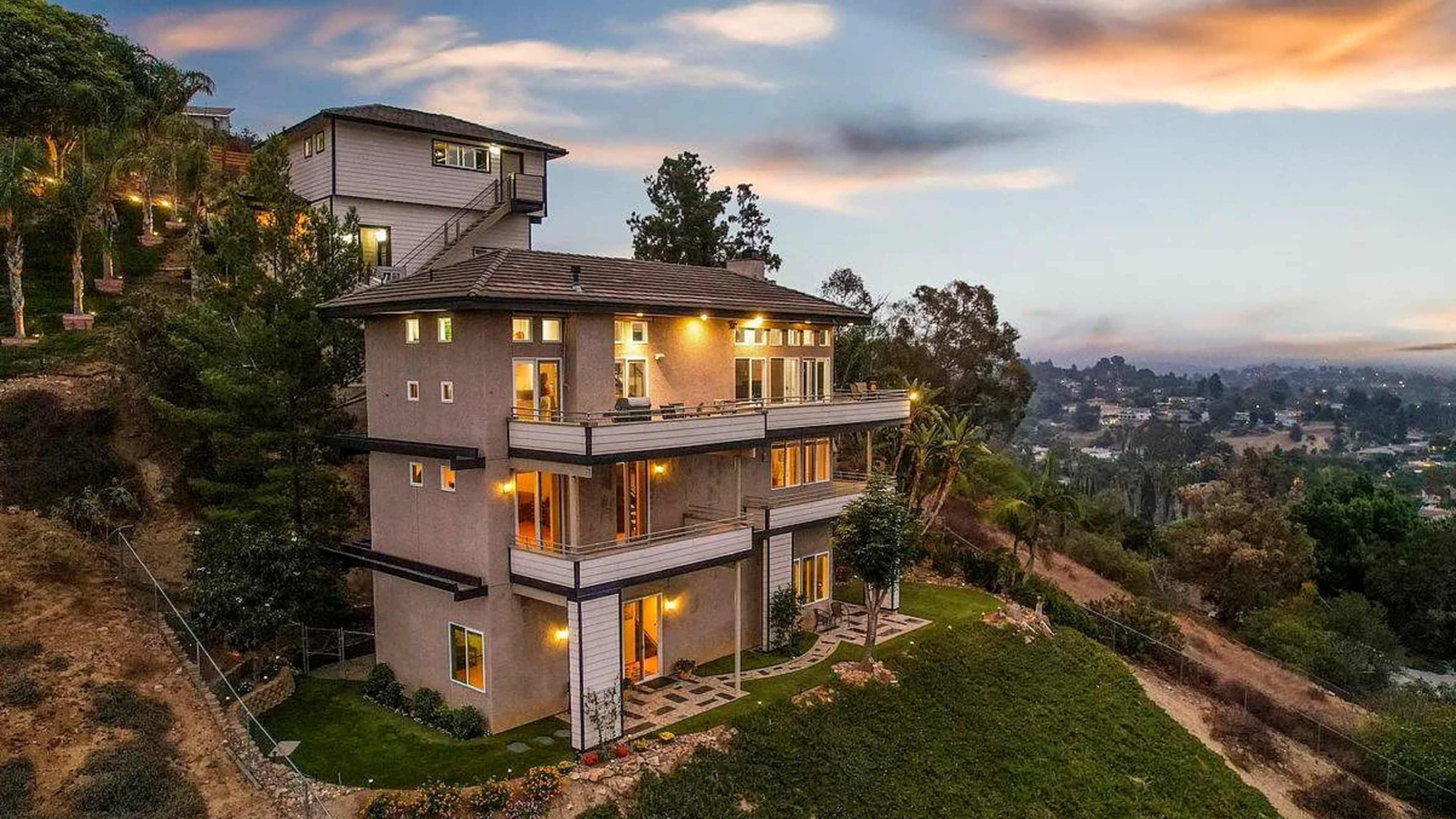 A modern multi-level house perched on a hillside, surrounded by trees and gardens, with lights glowing in the windows as dusk approaches.