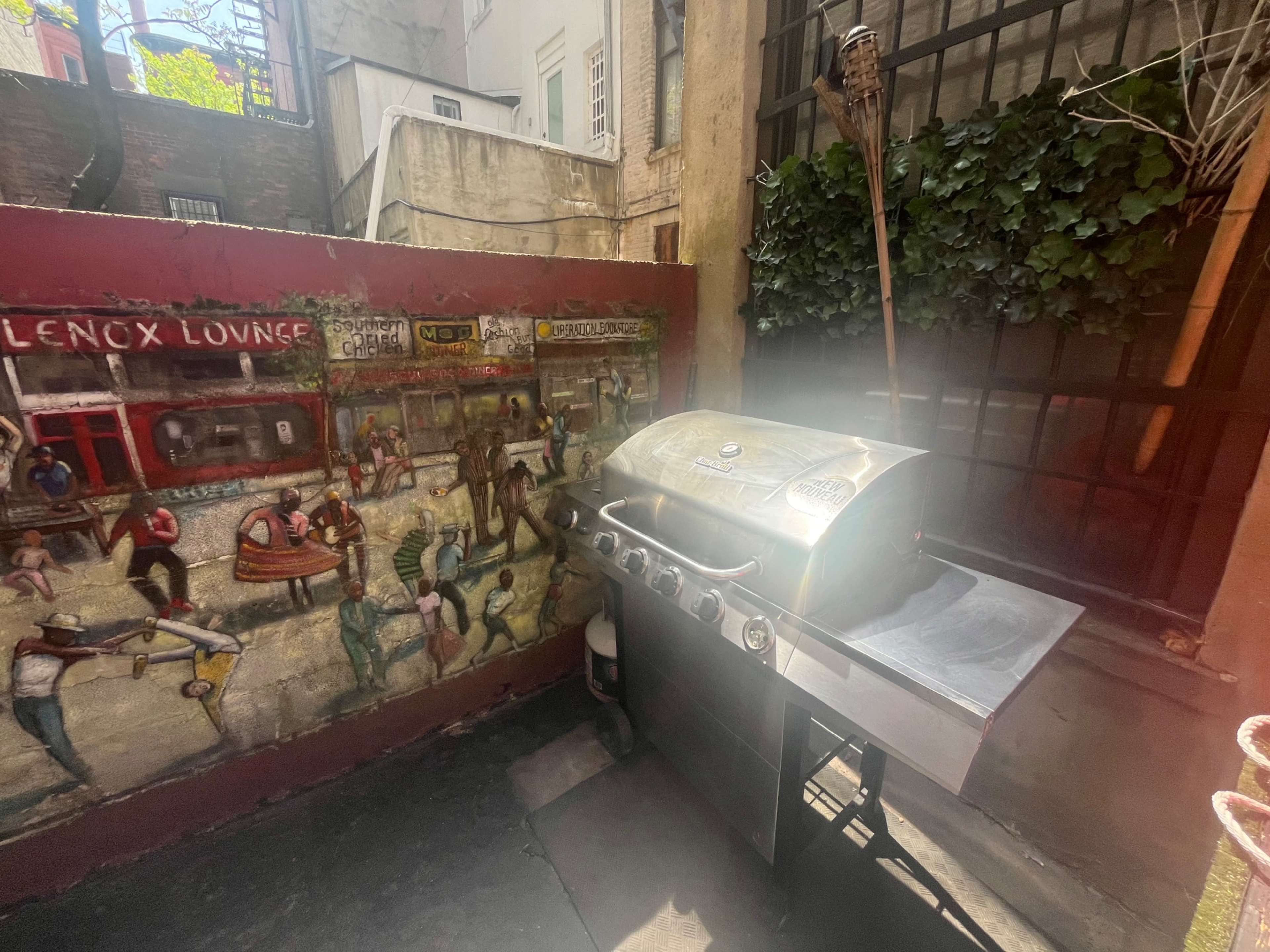 The image shows an outdoor grilling area featuring a stainless steel barbecue grill next to a colorful mural depicting people in a lively street scene.