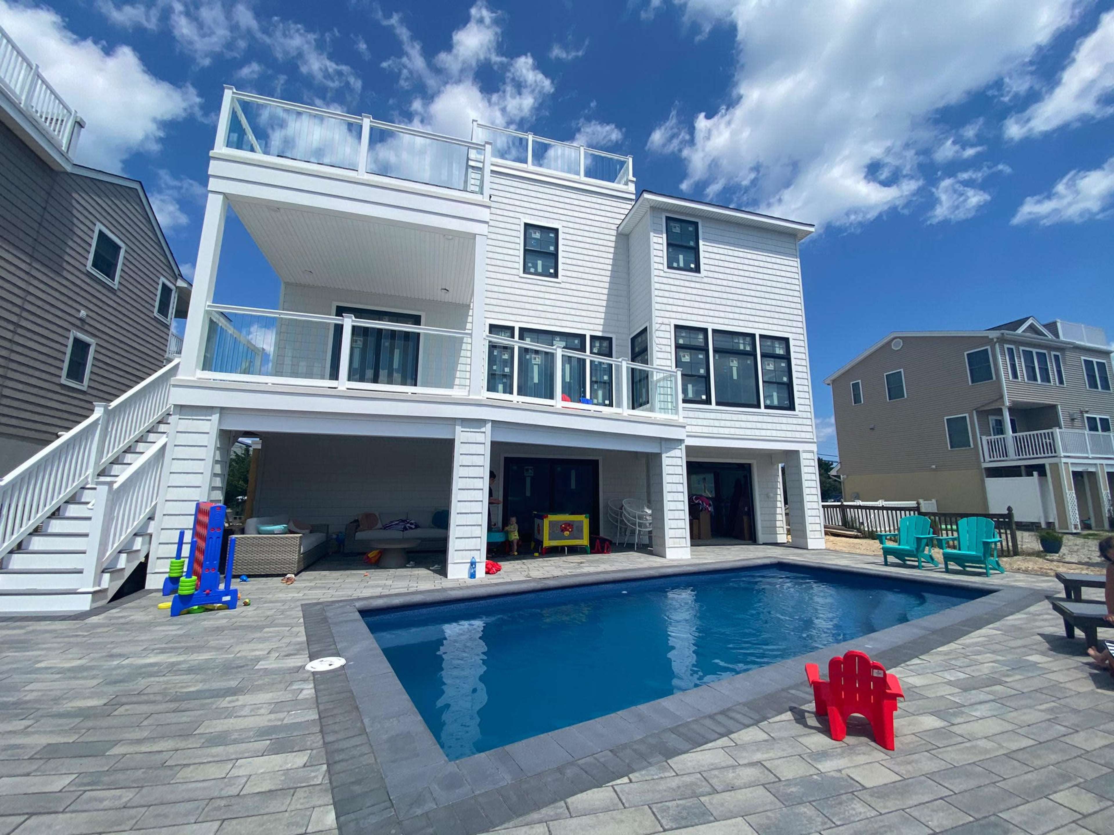 The image shows a two-story house with a spacious backyard featuring a swimming pool and colorful outdoor furniture under a partly cloudy sky.