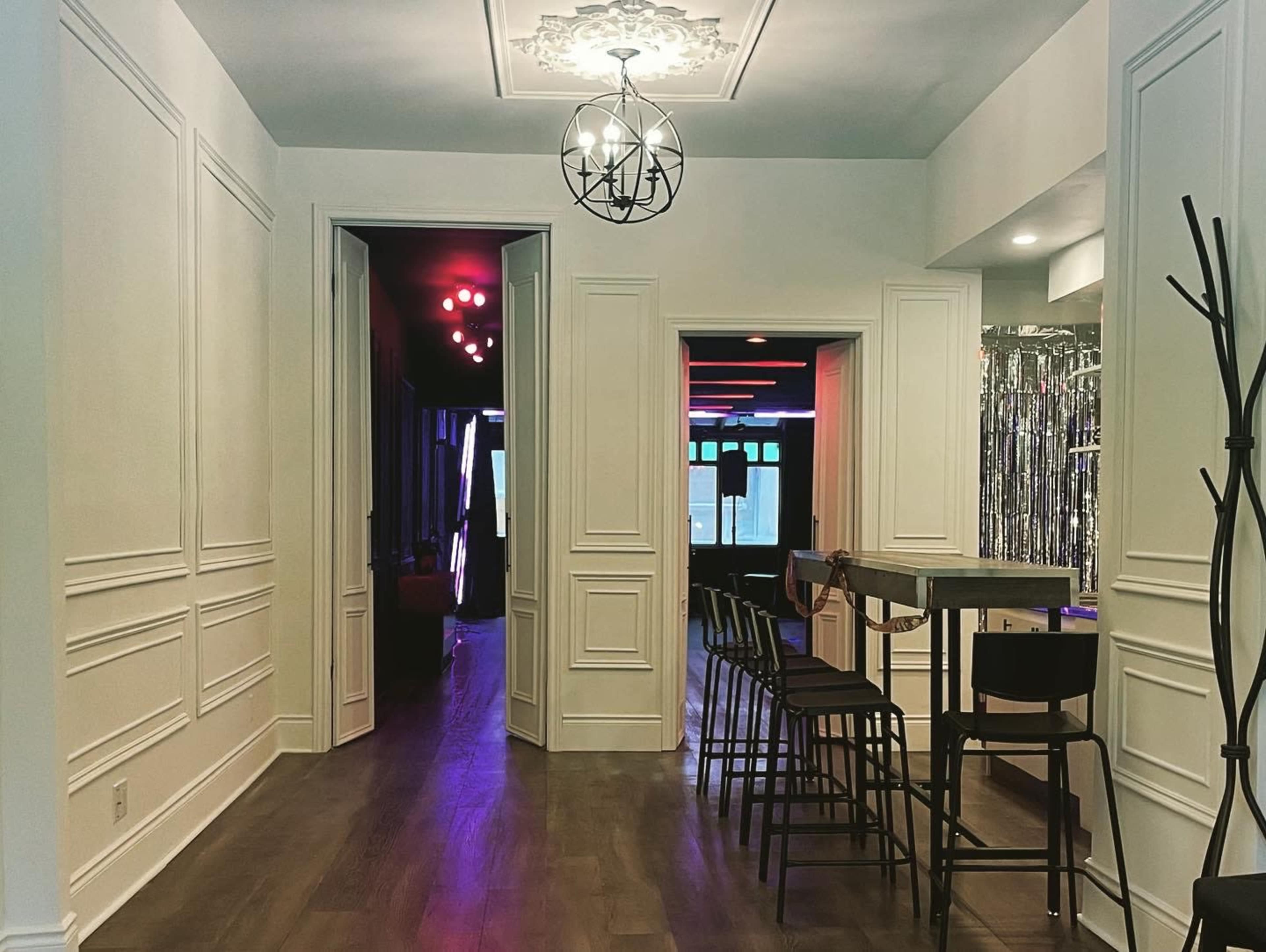 A modern hallway features white paneled walls, a chandelier overhead, and an open entrance to a brightly lit lounge area with a bar and seating.
