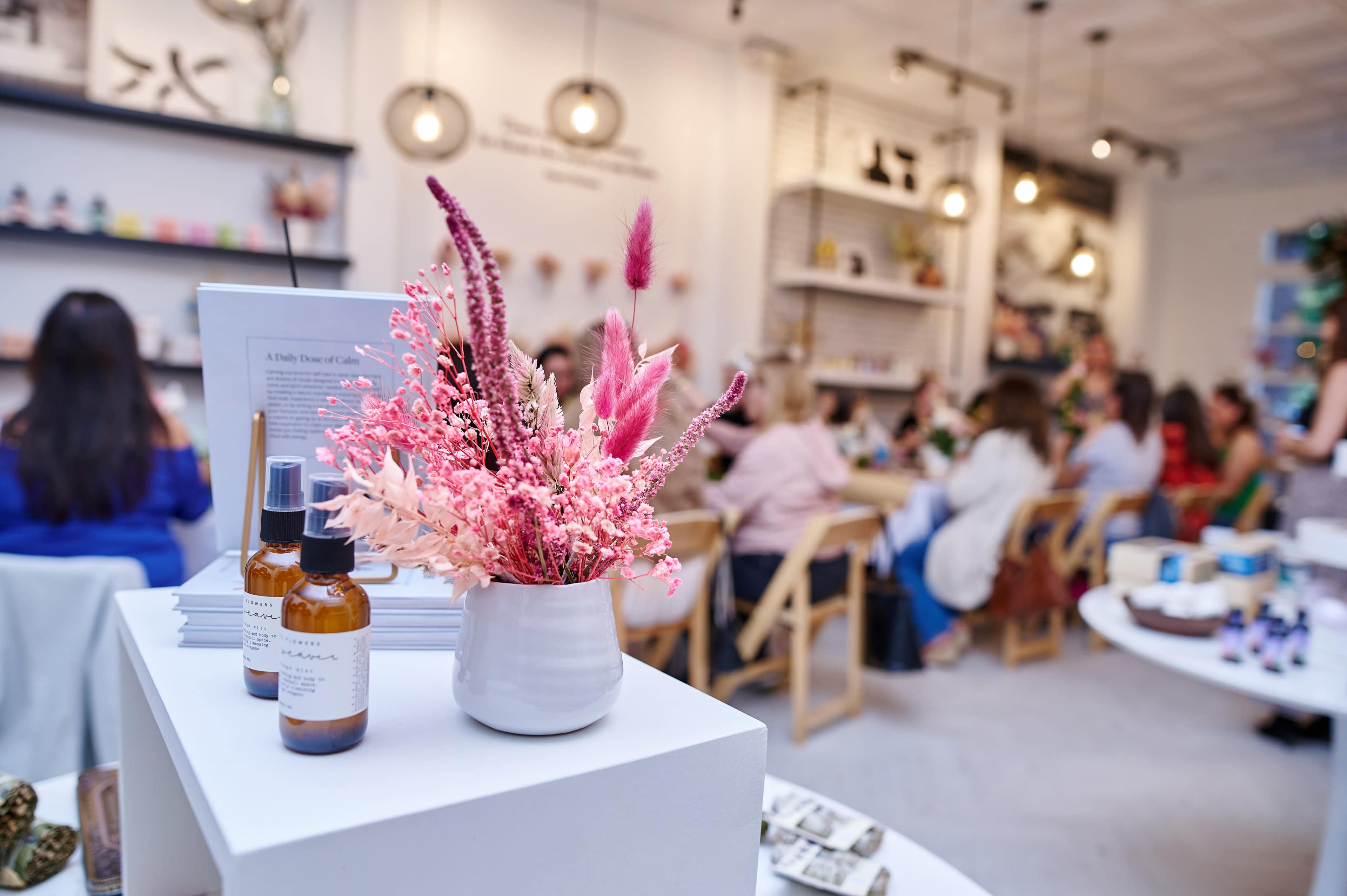 A cozy shop interior with a display of skincare products and a flower arrangement in a vase, while customers engage in conversation at tables in the background.