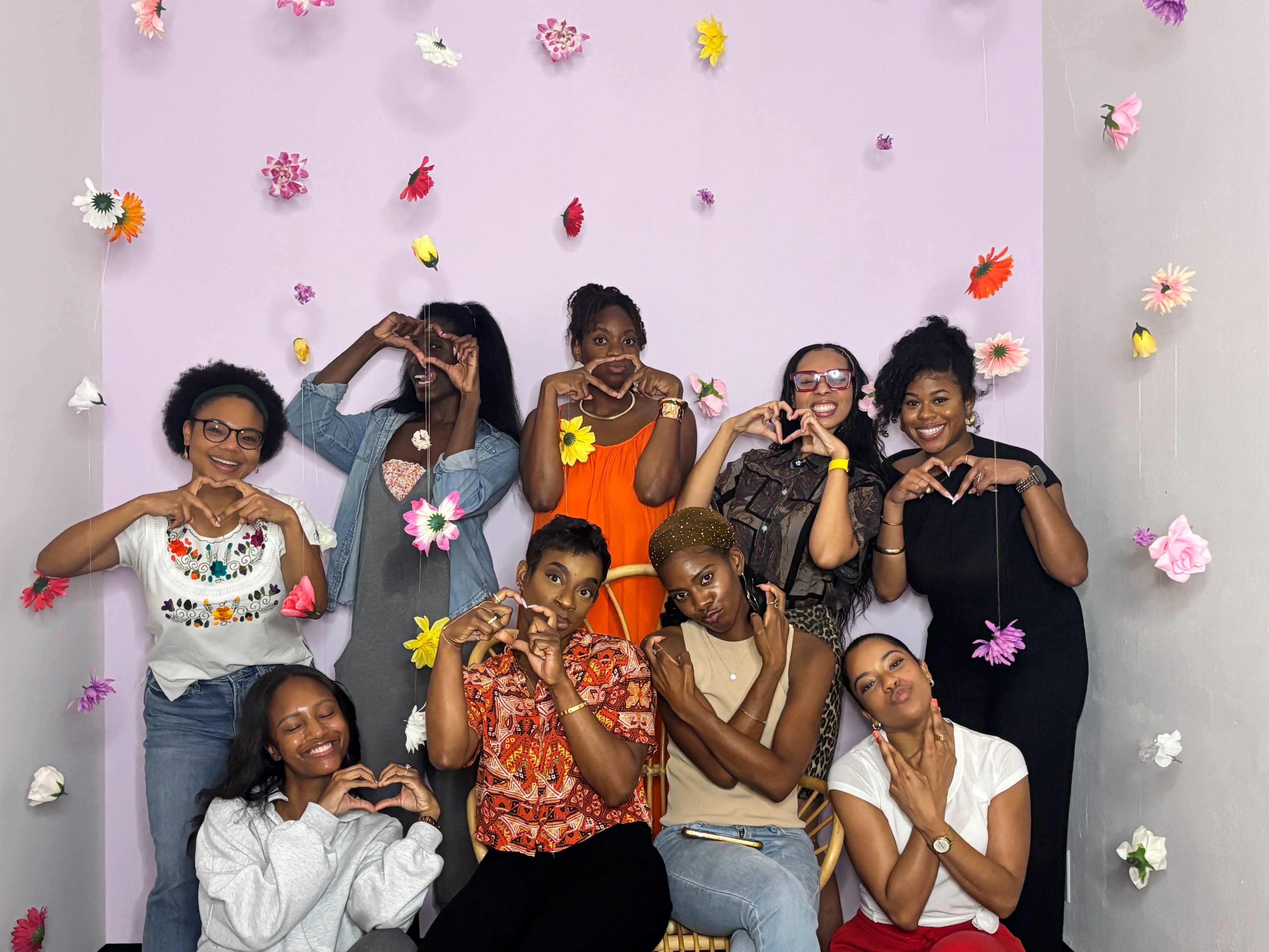 A group of nine women poses in front of a pink wall adorned with colorful paper flowers, each making heart shapes with their hands.