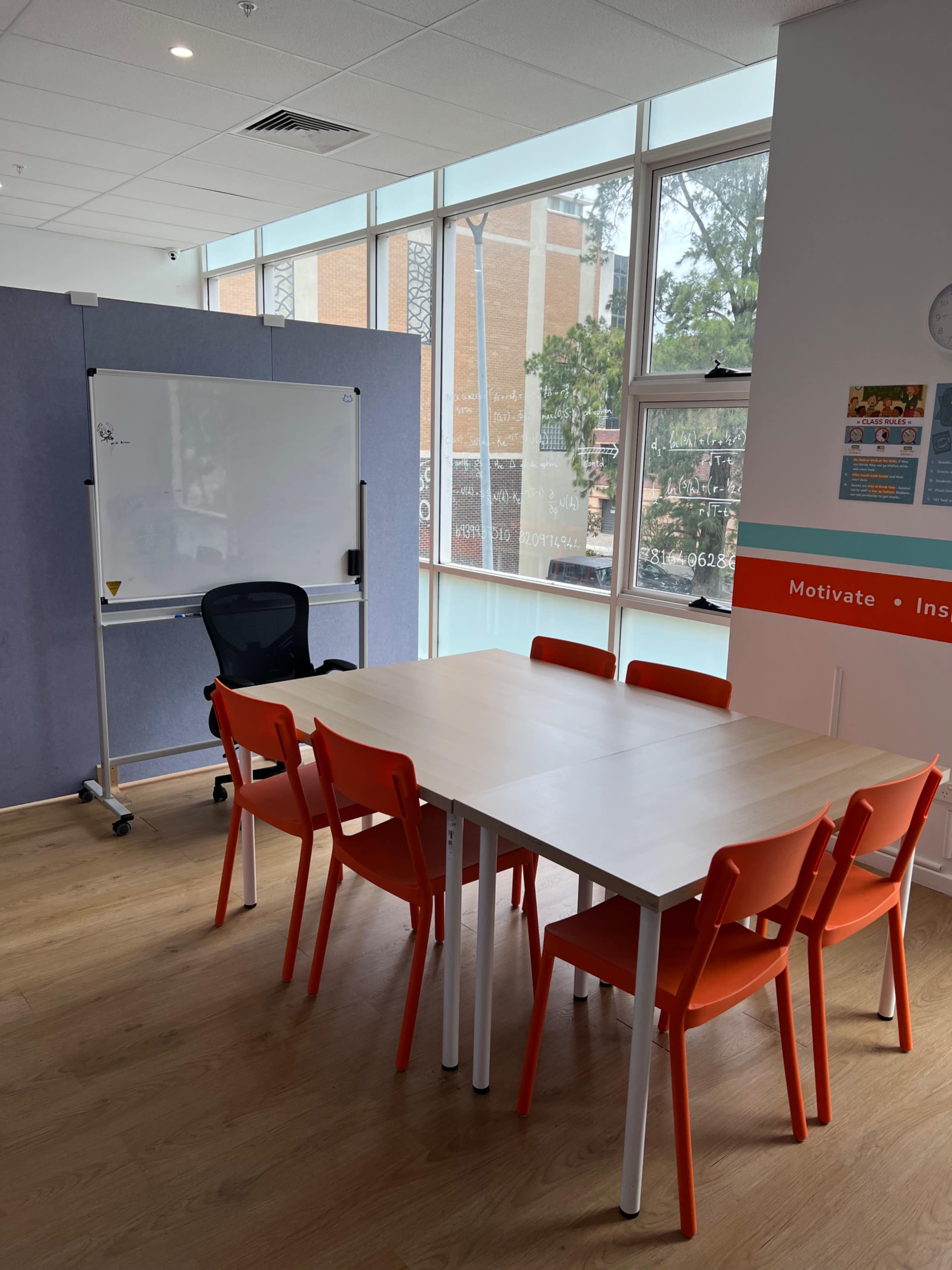 A meeting room features a large table surrounded by orange chairs, a whiteboard on wheels, and large windows allowing natural light.