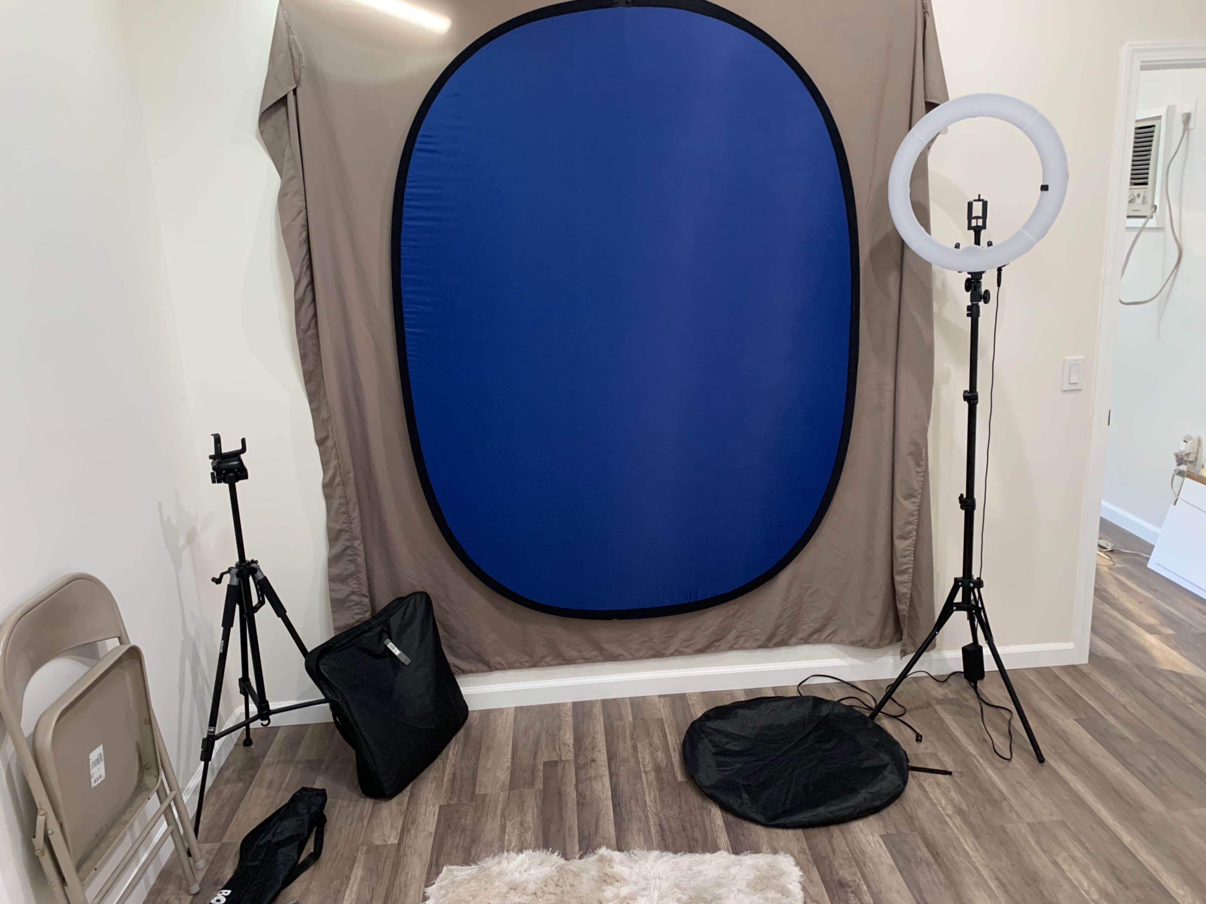 The image shows a photography setup featuring a blue backdrop, a ring light on a stand, and tripods in a room with hardwood flooring.