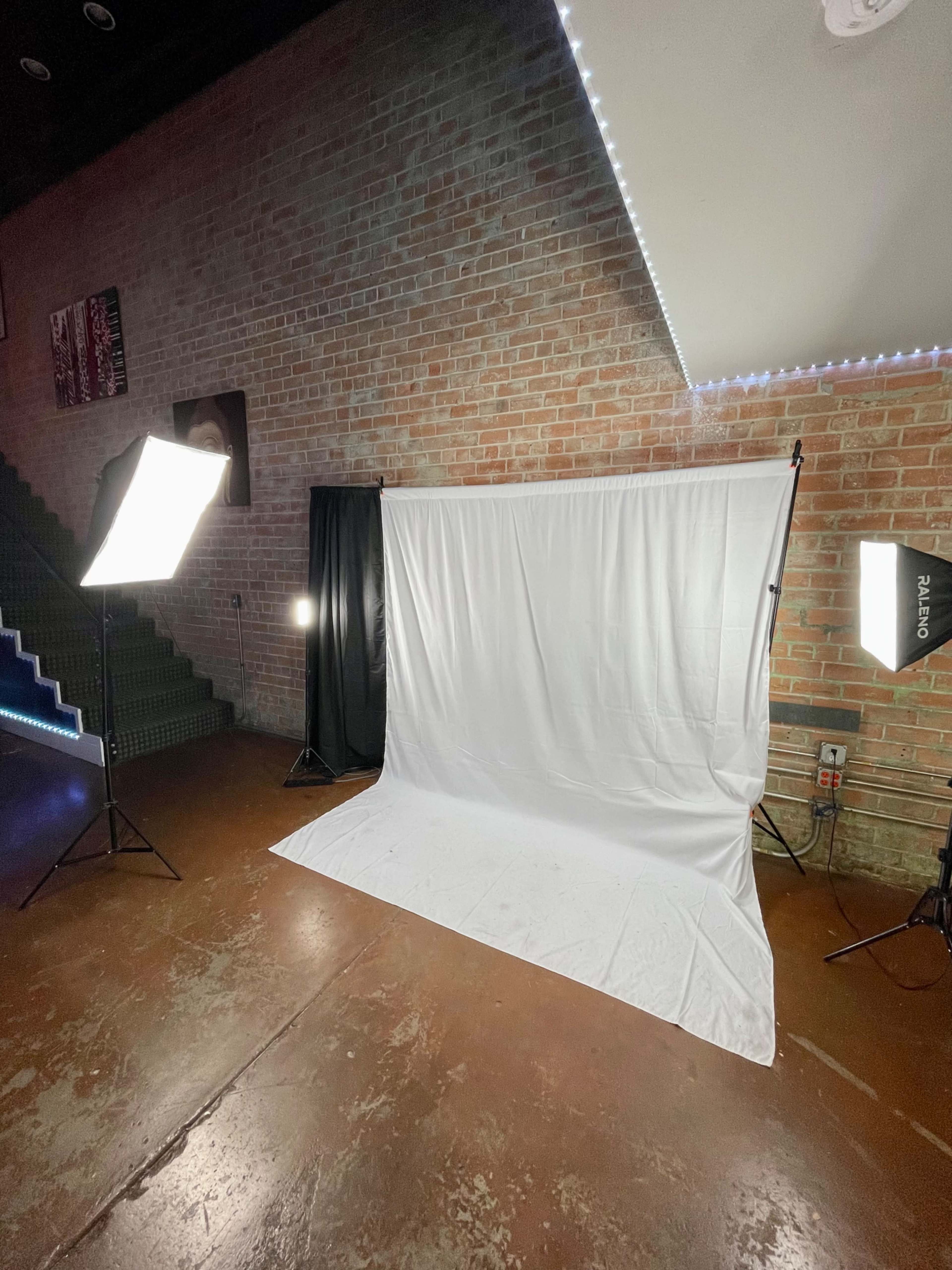 A spacious room features a white backdrop with two bright lights positioned on stands nearby, alongside a staircase in the background.