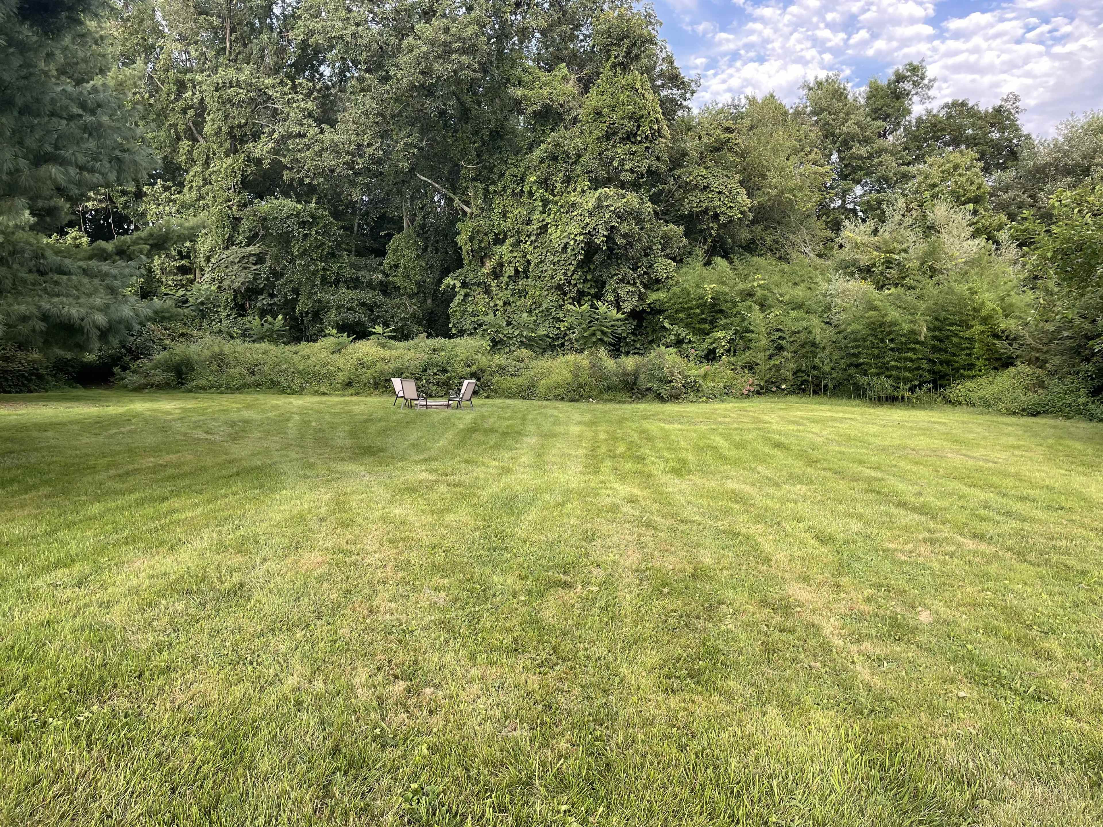 A wide, grassy field features two empty chairs positioned in the center, surrounded by dense greenery and trees.