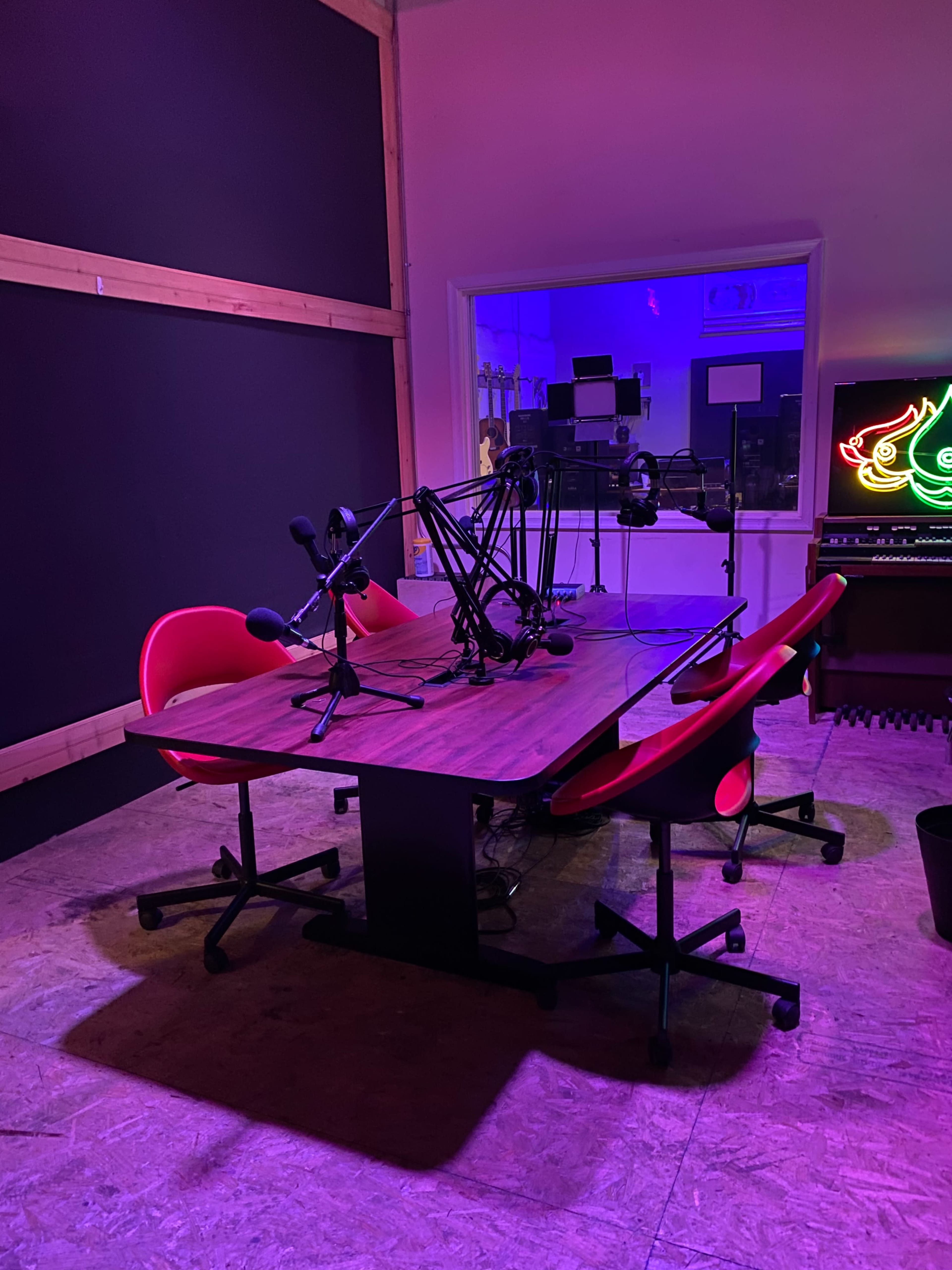 A podcast studio featuring a wooden table with microphones and four red chairs arranged around it, illuminated by purple and blue lighting.