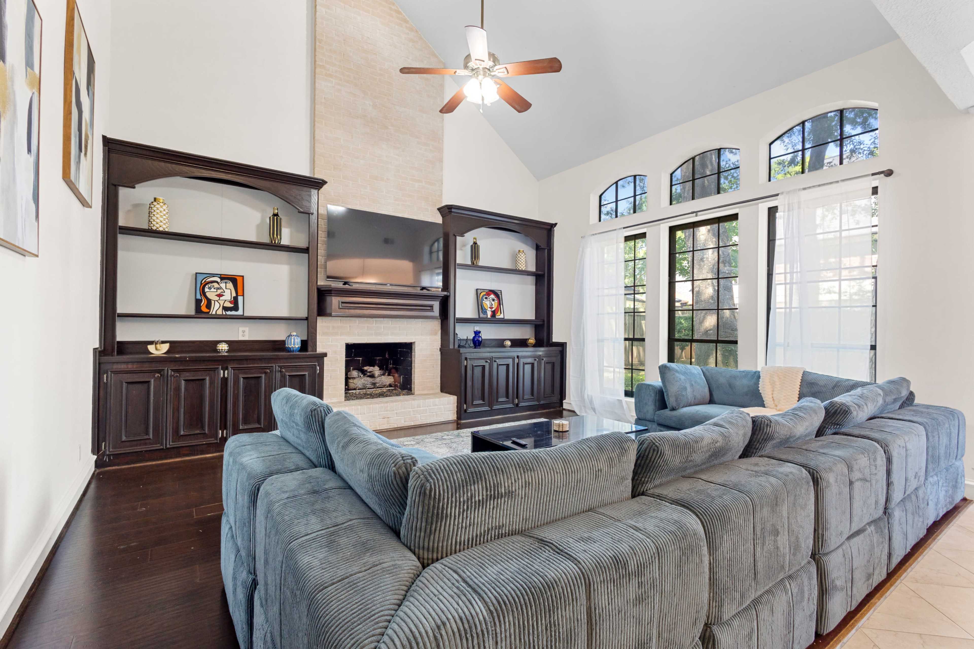 A spacious living room features a large, gray sectional sofa facing a dark wooden entertainment center with a fireplace and large windows providing natural light.
