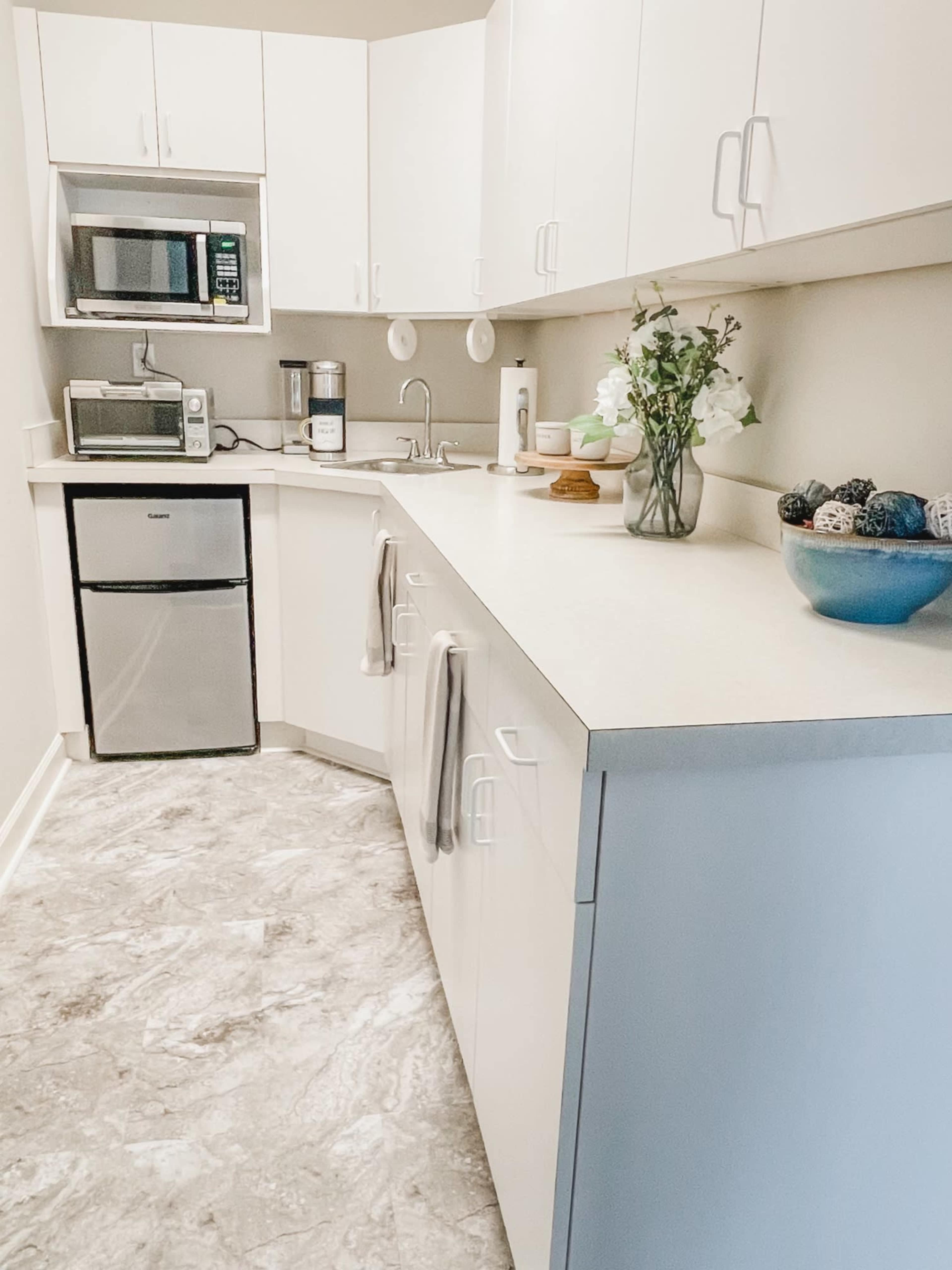 A compact kitchen features white cabinets, a stainless steel microwave, a mini refrigerator, and a countertop adorned with a vase of flowers and decorative items.