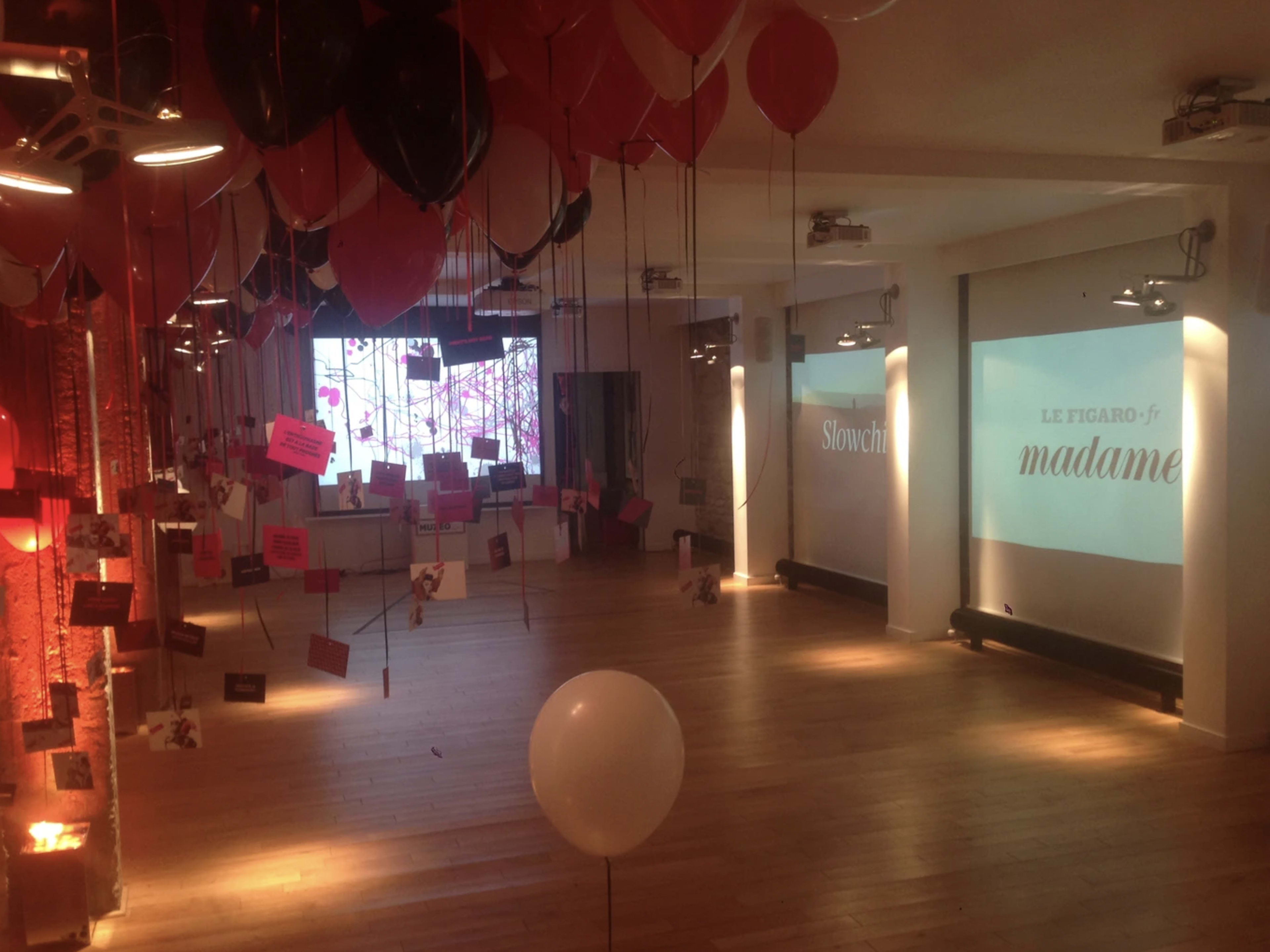 A large indoor space is decorated with red and black balloons, string lights, and hanging signs, featuring two projection screens on opposite walls.