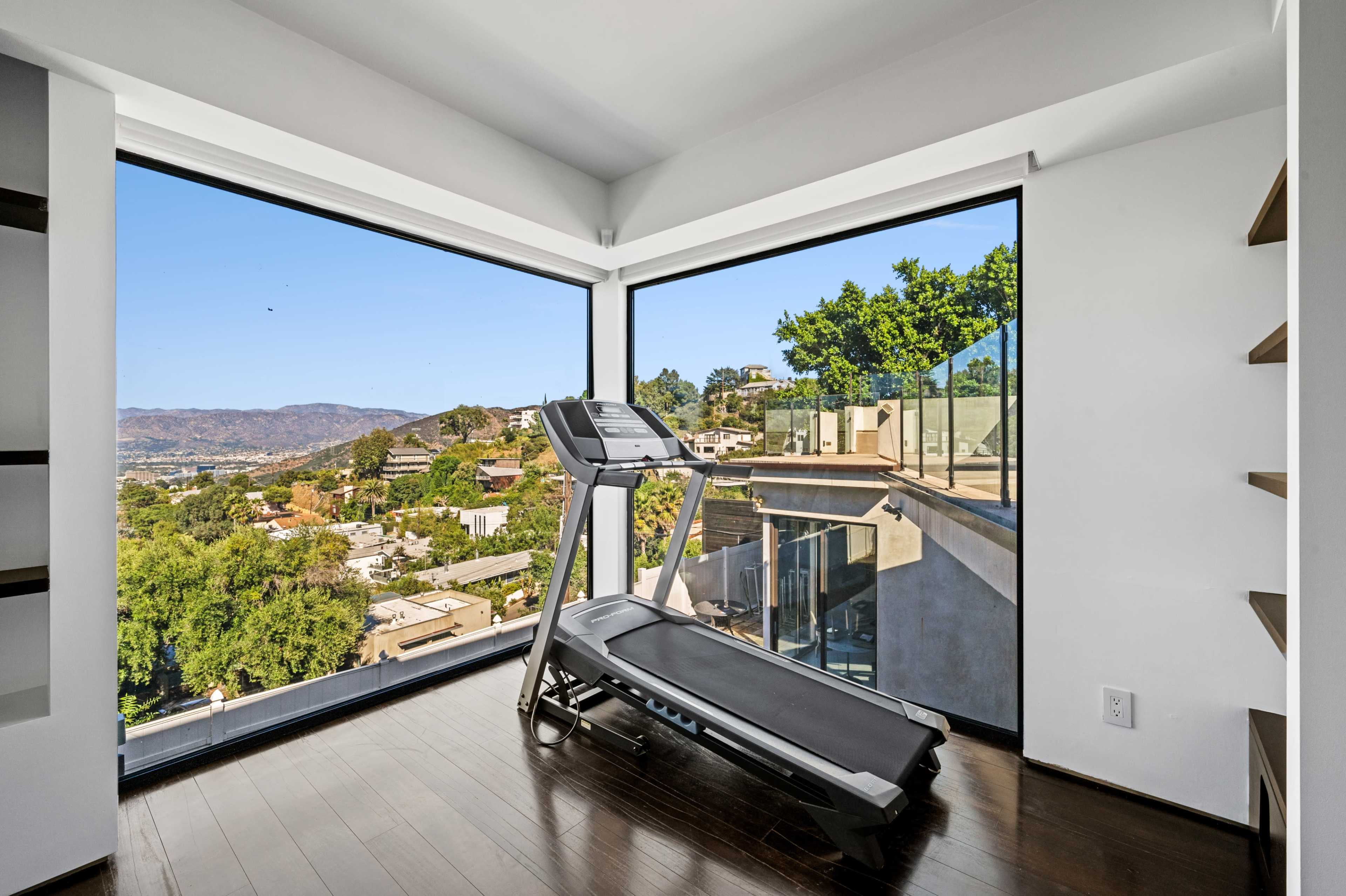 A treadmill is positioned in a room with large windows offering a view of a hilly landscape and distant homes.