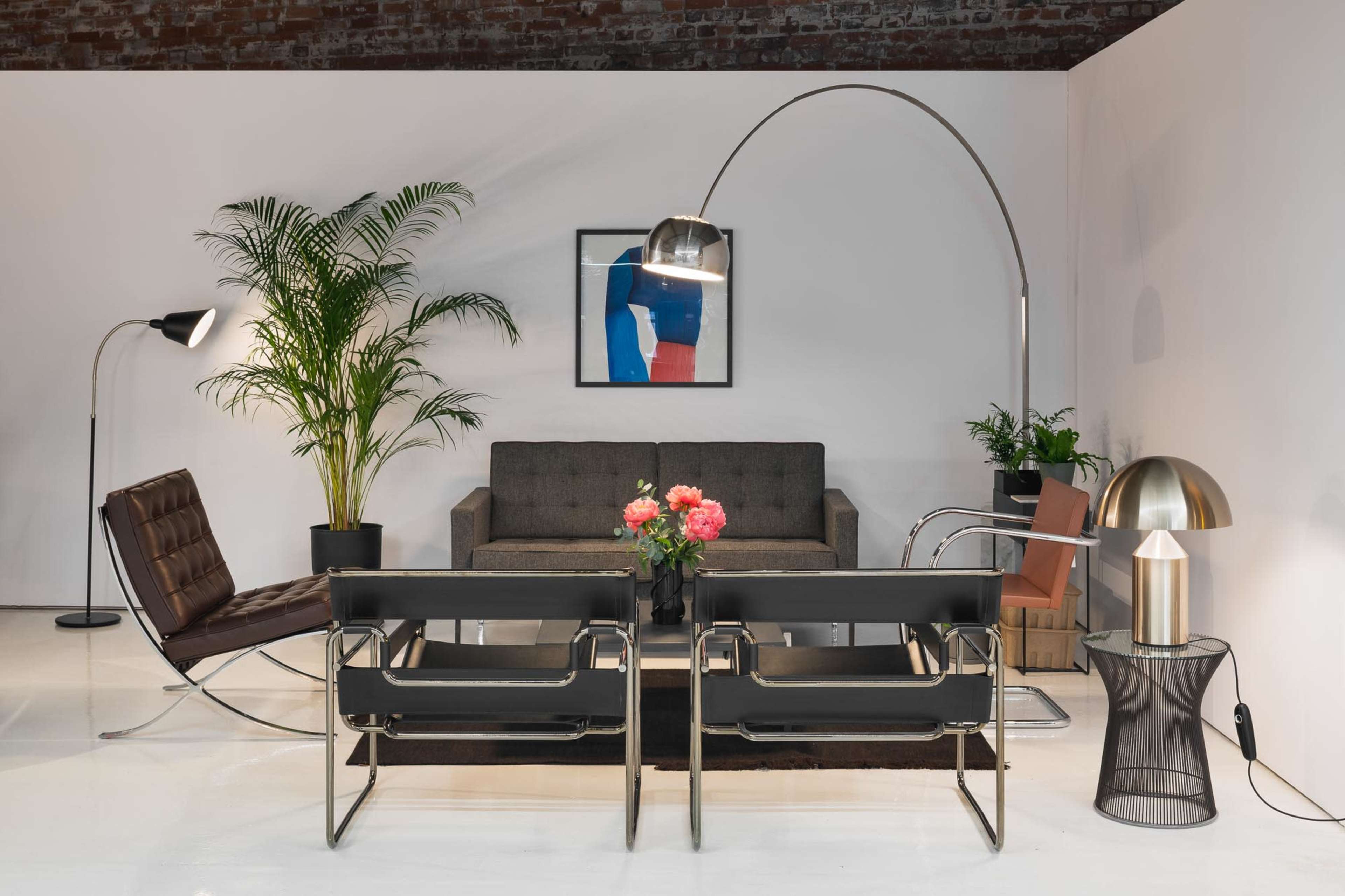 The image shows a modern interior with a gray sofa, two black chairs, a floor lamp, a table lamp, and a potted plant against a white wall.