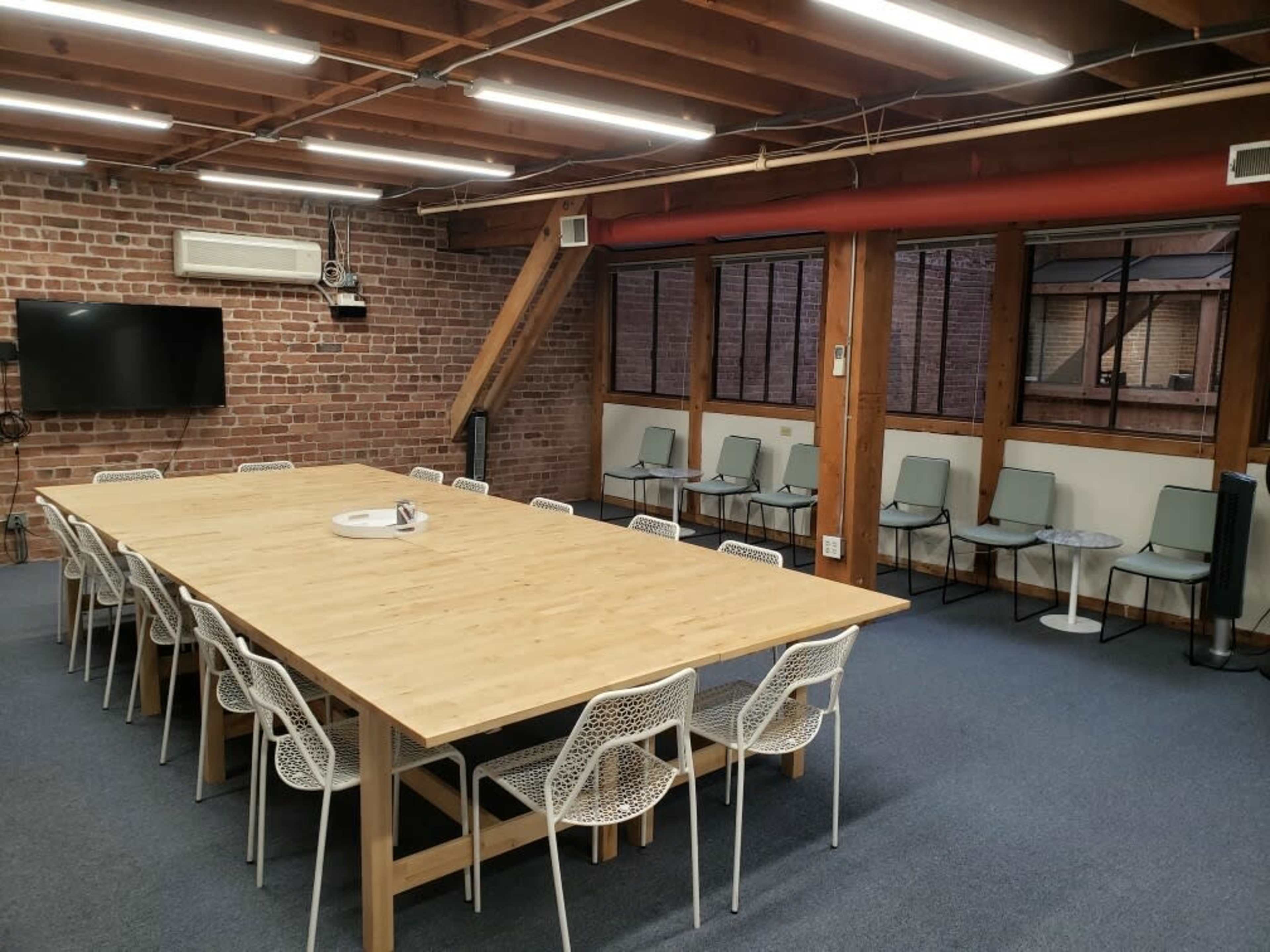 A large wooden table surrounded by white chairs occupies the center of a meeting room with exposed brick walls and large windows.