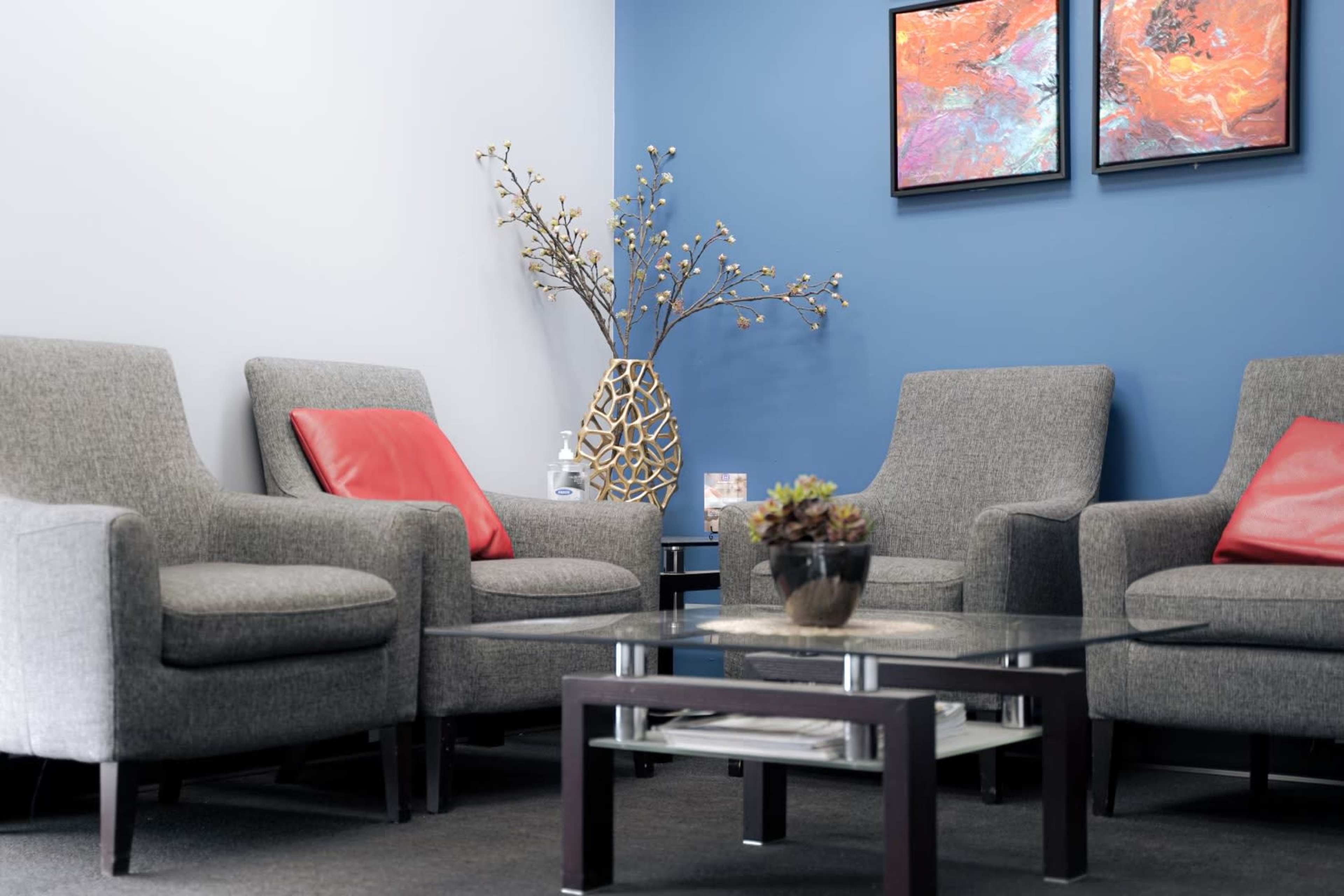 The image shows a waiting area with two grey armchairs featuring red cushions, a glass coffee table, and a decorative vase with branches against a blue wall.