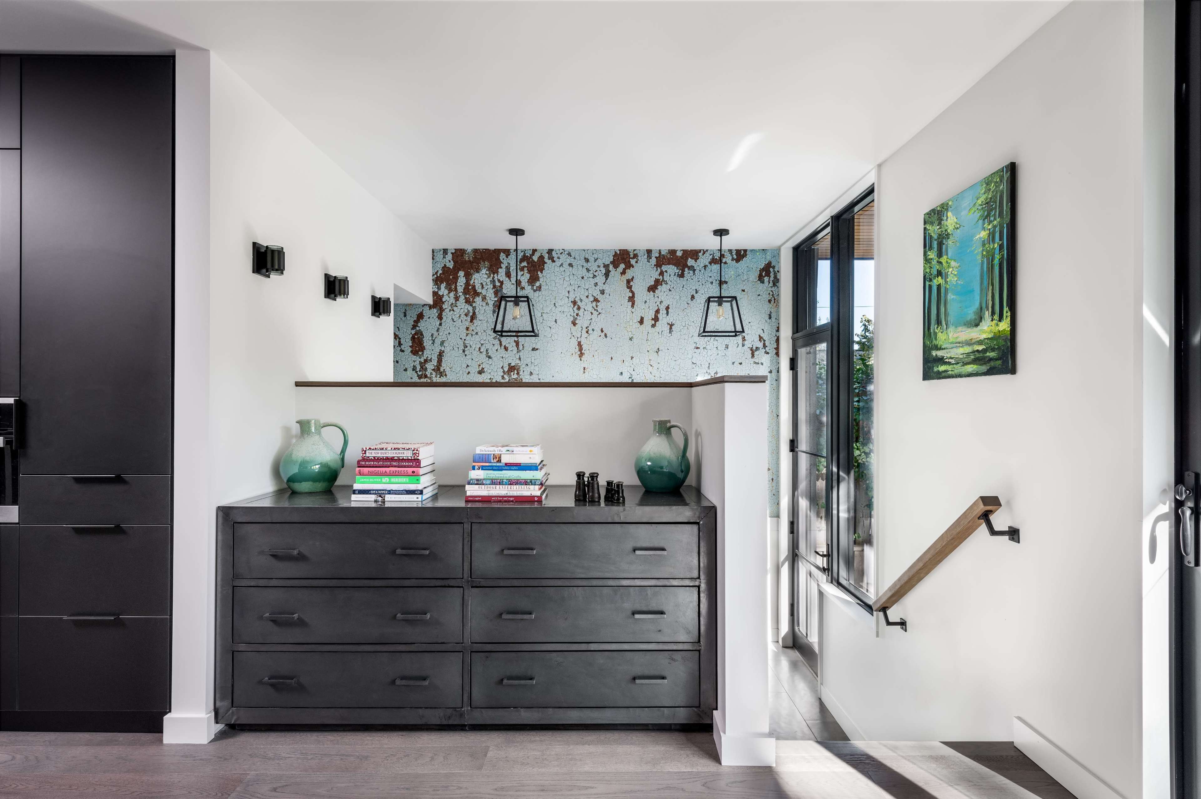 A modern entryway with a gray dresser topped with stacked magazines and decorative vases, adjacent to a light-filled space featuring a colorful, textured wall and a staircase.