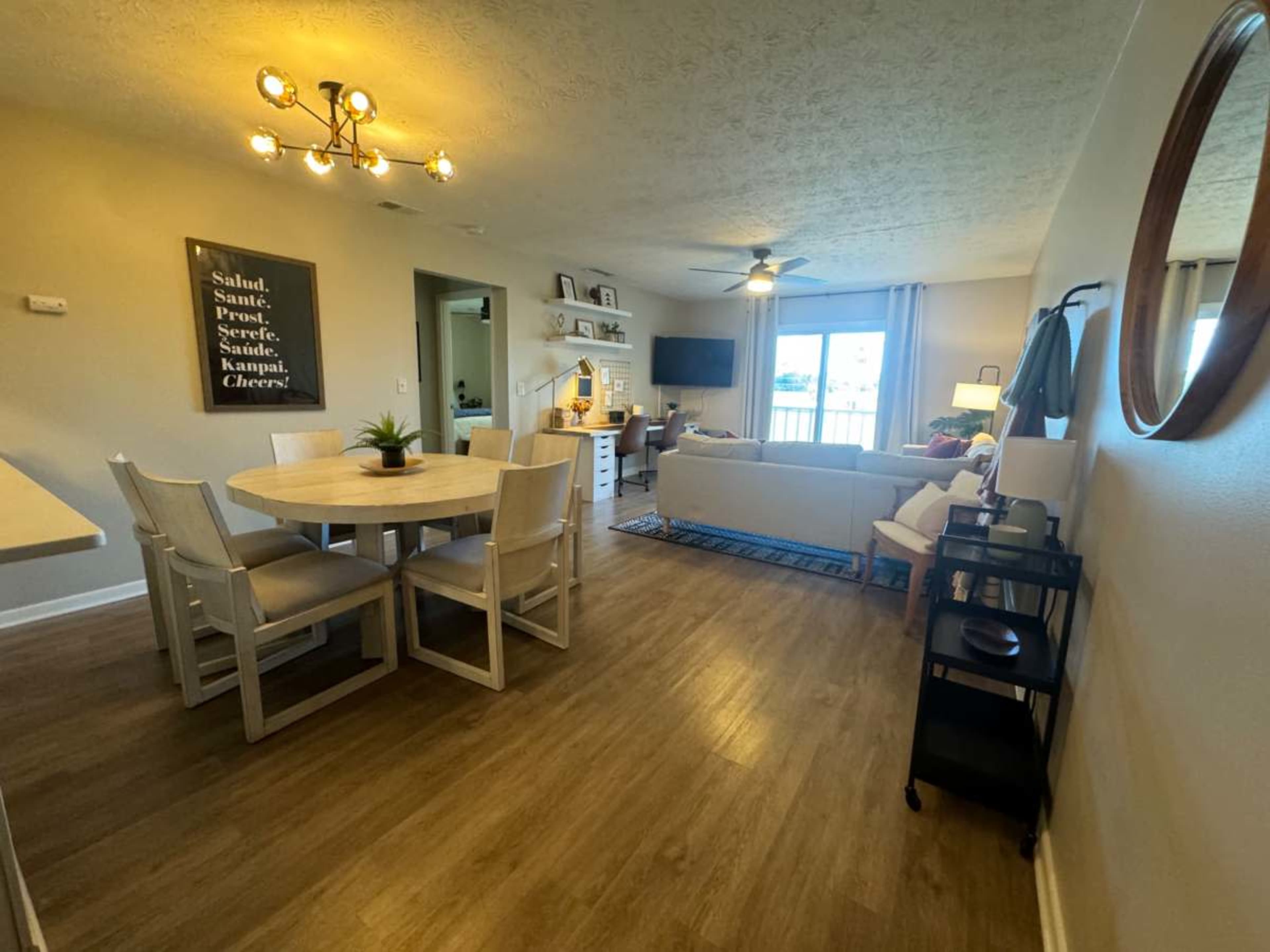 The image shows a bright living area featuring a circular dining table, a beige sofa, a television on a stand, and decorative elements on the walls.