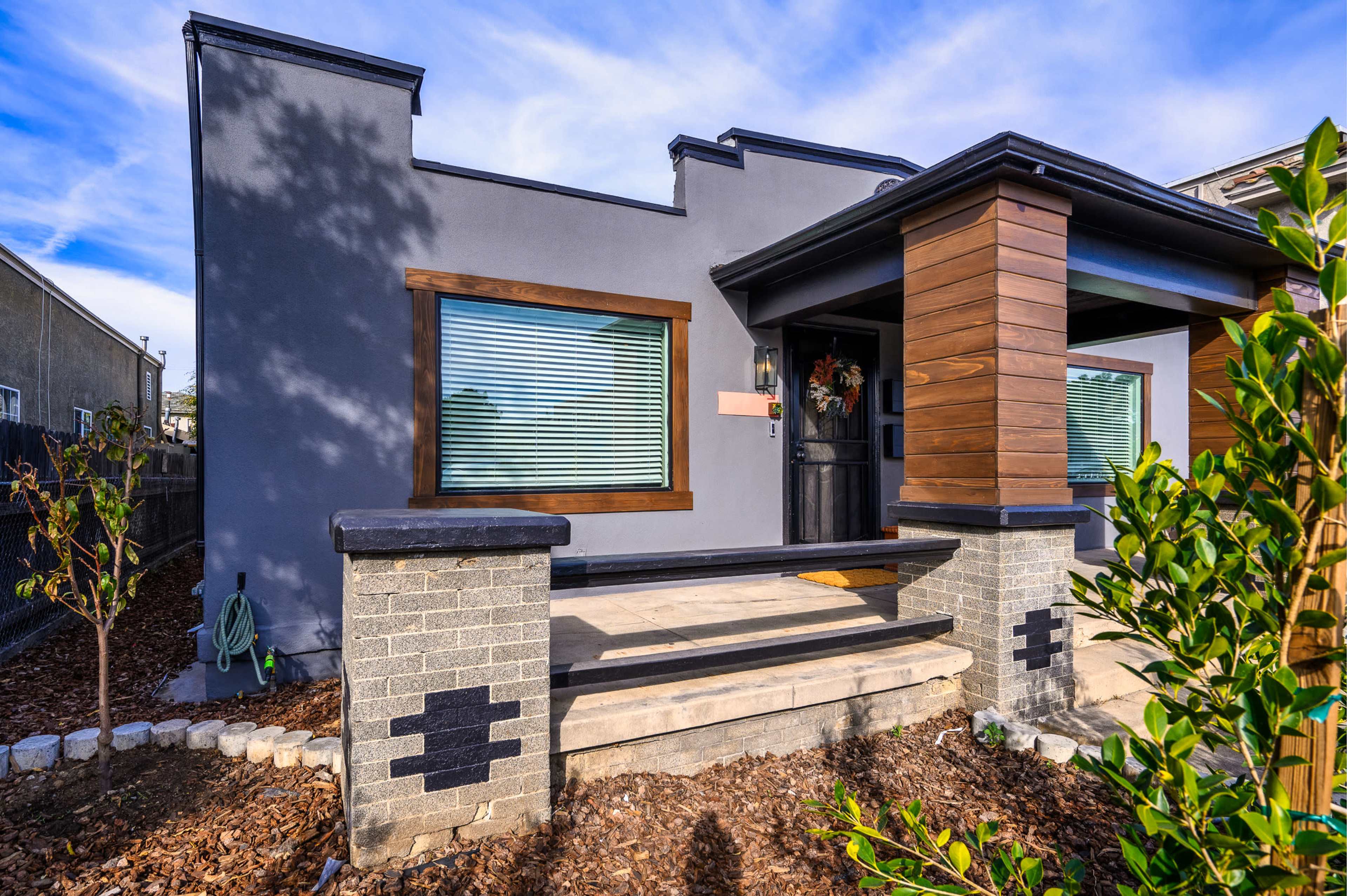 The image shows a modern single-story house with a gray exterior and wooden accents, featuring a stone front porch and landscaped garden.