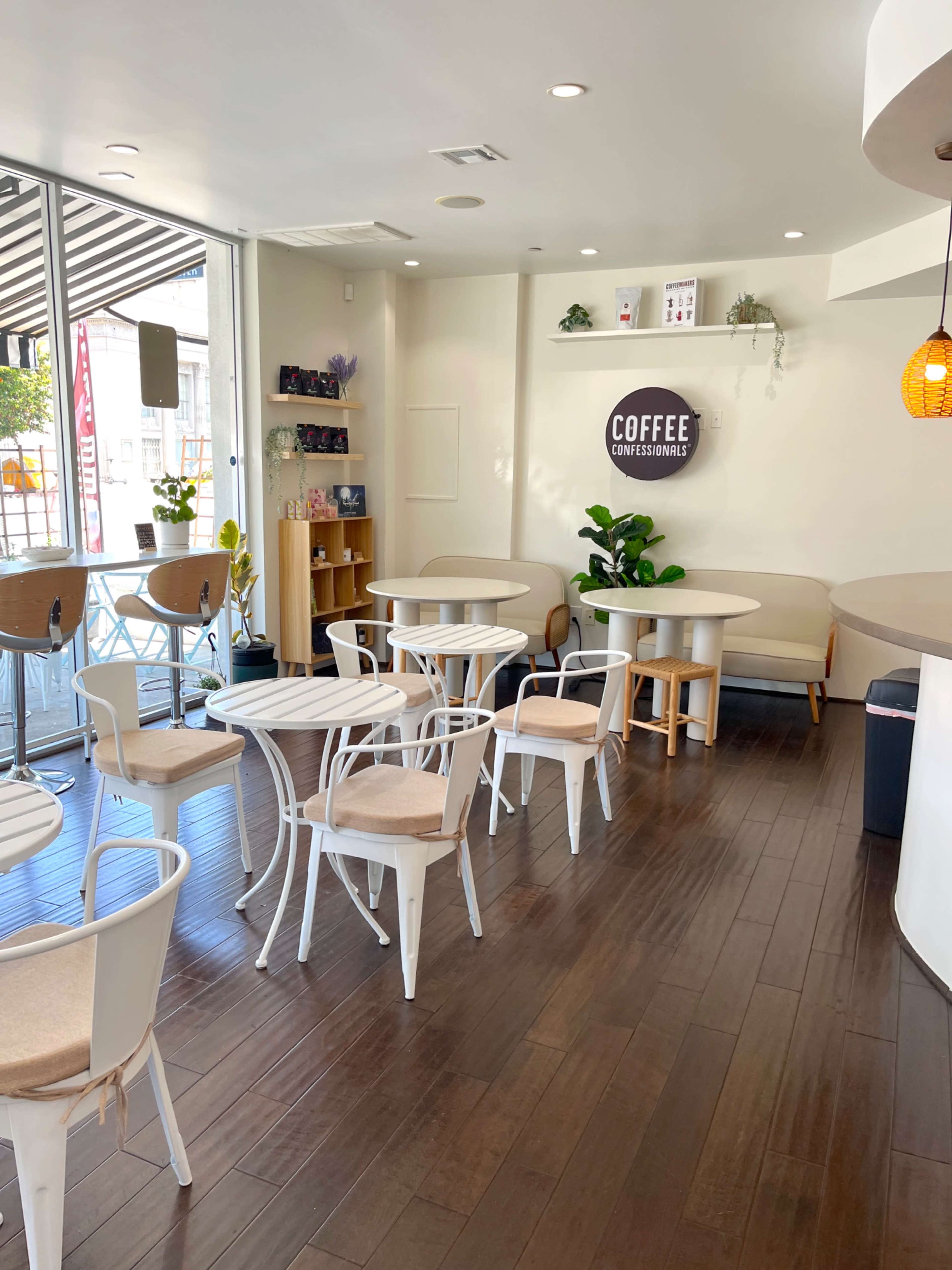 The image shows a modern coffee shop with white furniture, wooden flooring, and large windows allowing natural light to enter.