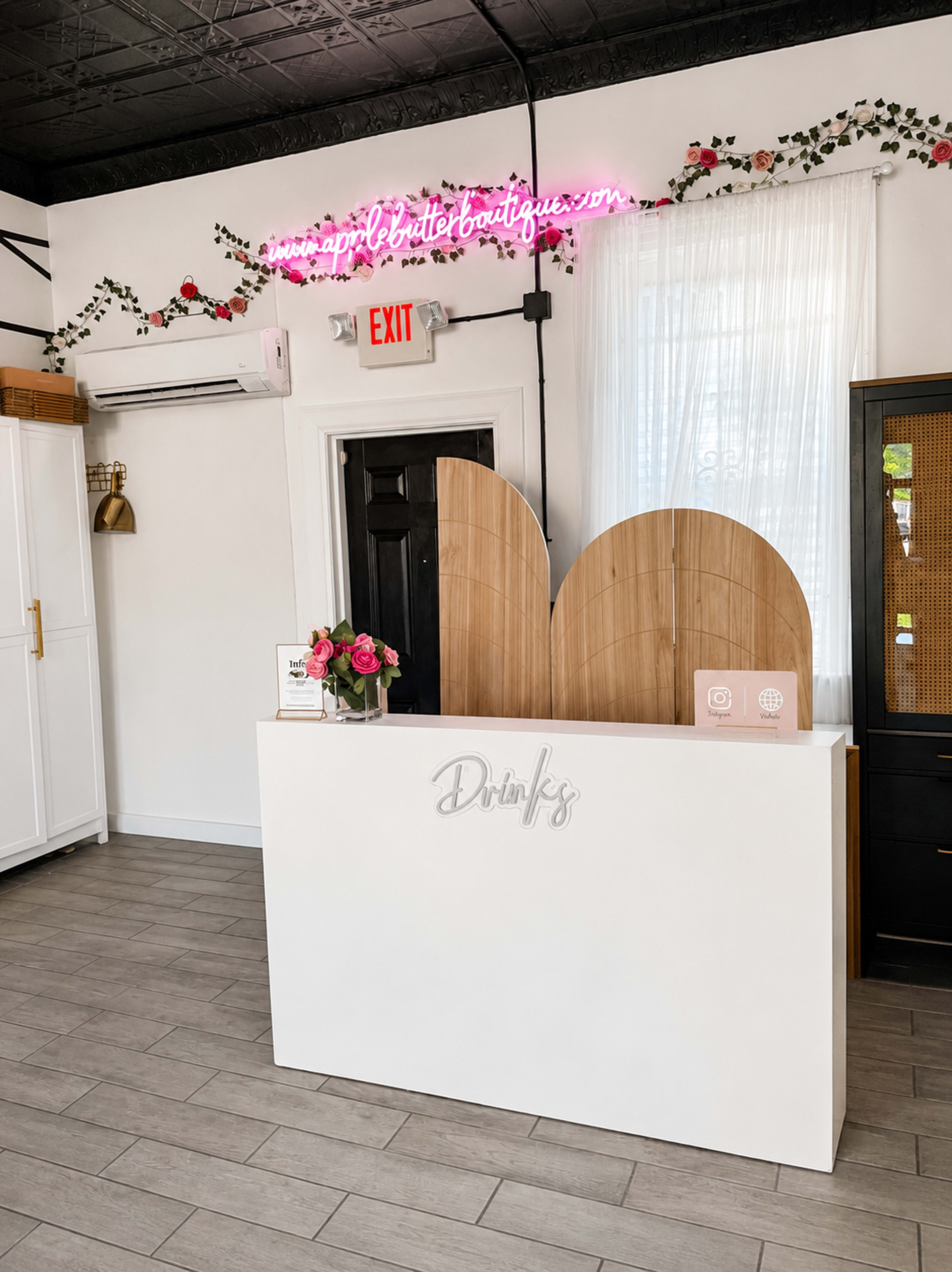 The image features a boutique reception area with a white counter, floral decorations on the wall, and a neon sign that reads "Drinks."