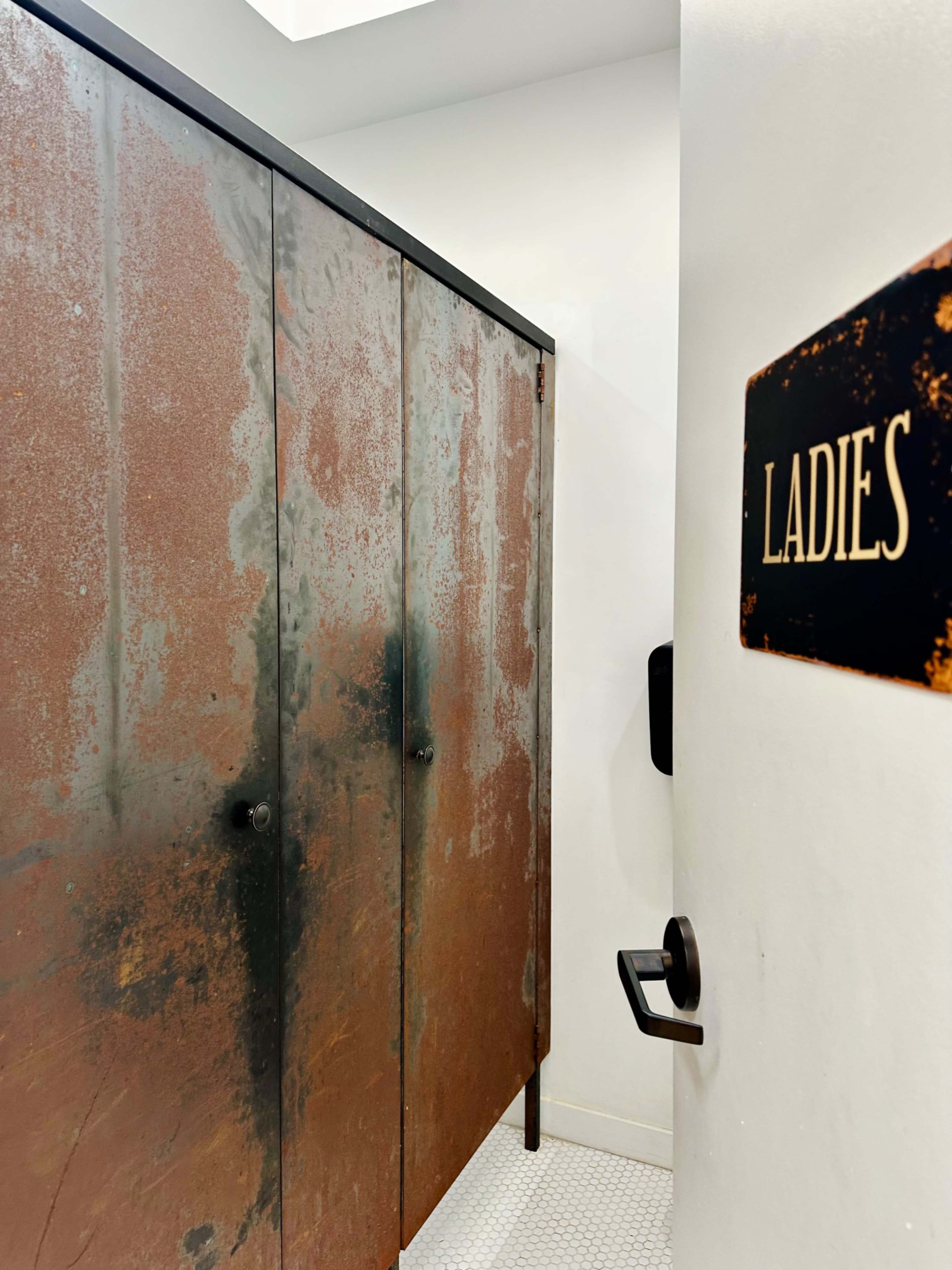 The image shows a restroom entrance labeled "LADIES" next to a set of rusted metal stall doors.