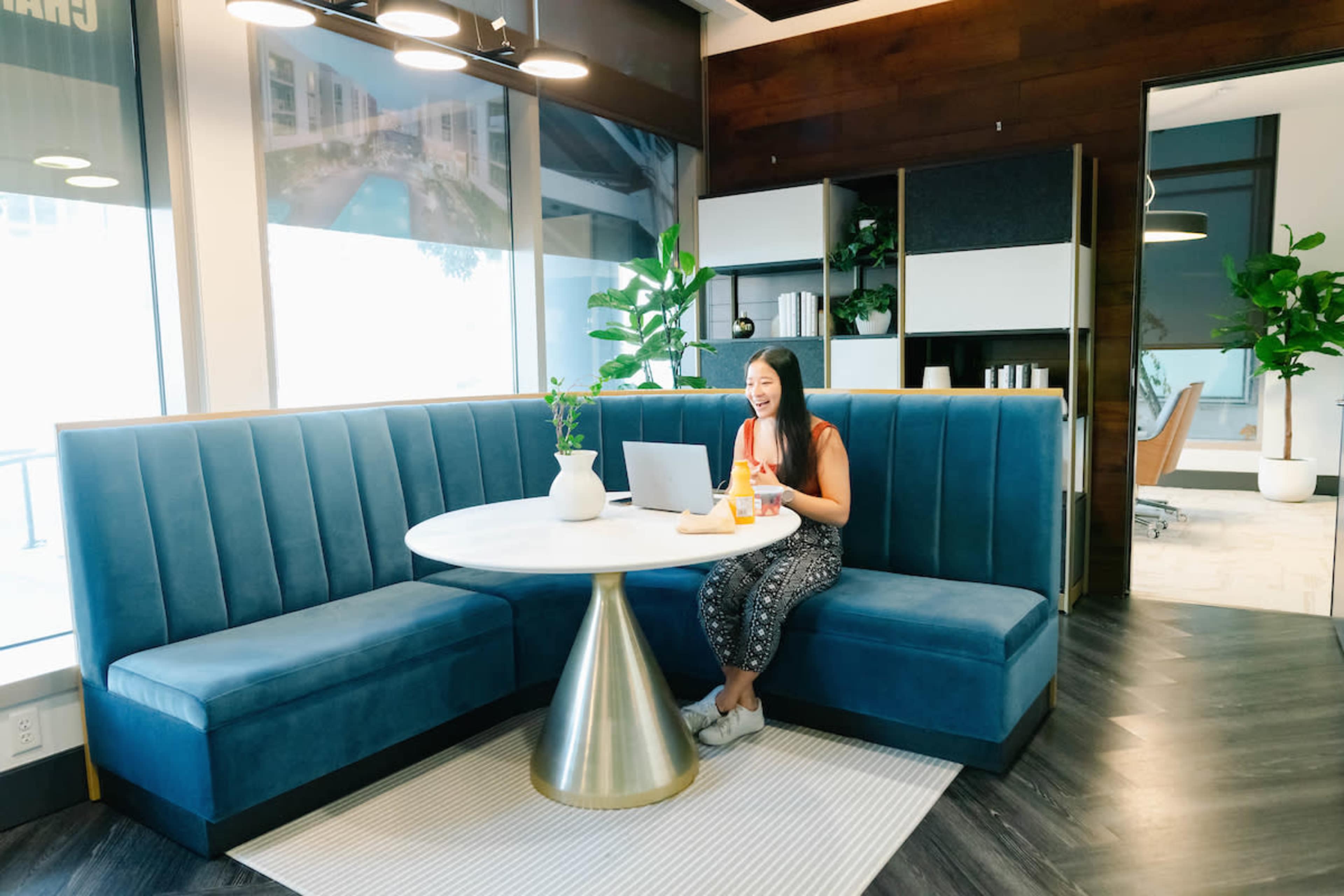A woman sits at a round table in a modern office space, working on a laptop with snacks beside her.