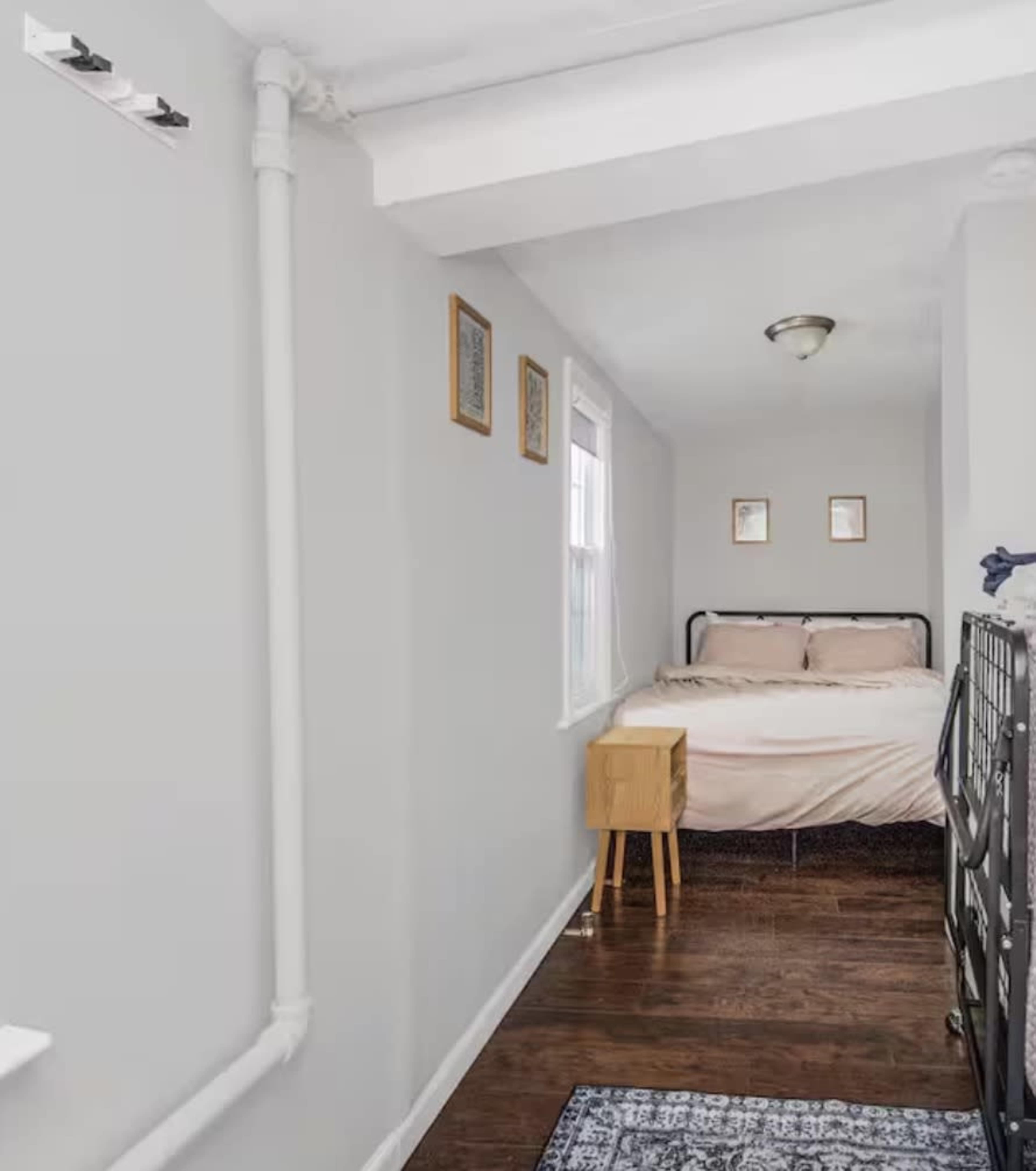 The image shows a narrow hallway leading to a small bedroom with a bed, wooden nightstand, and framed pictures on the walls.