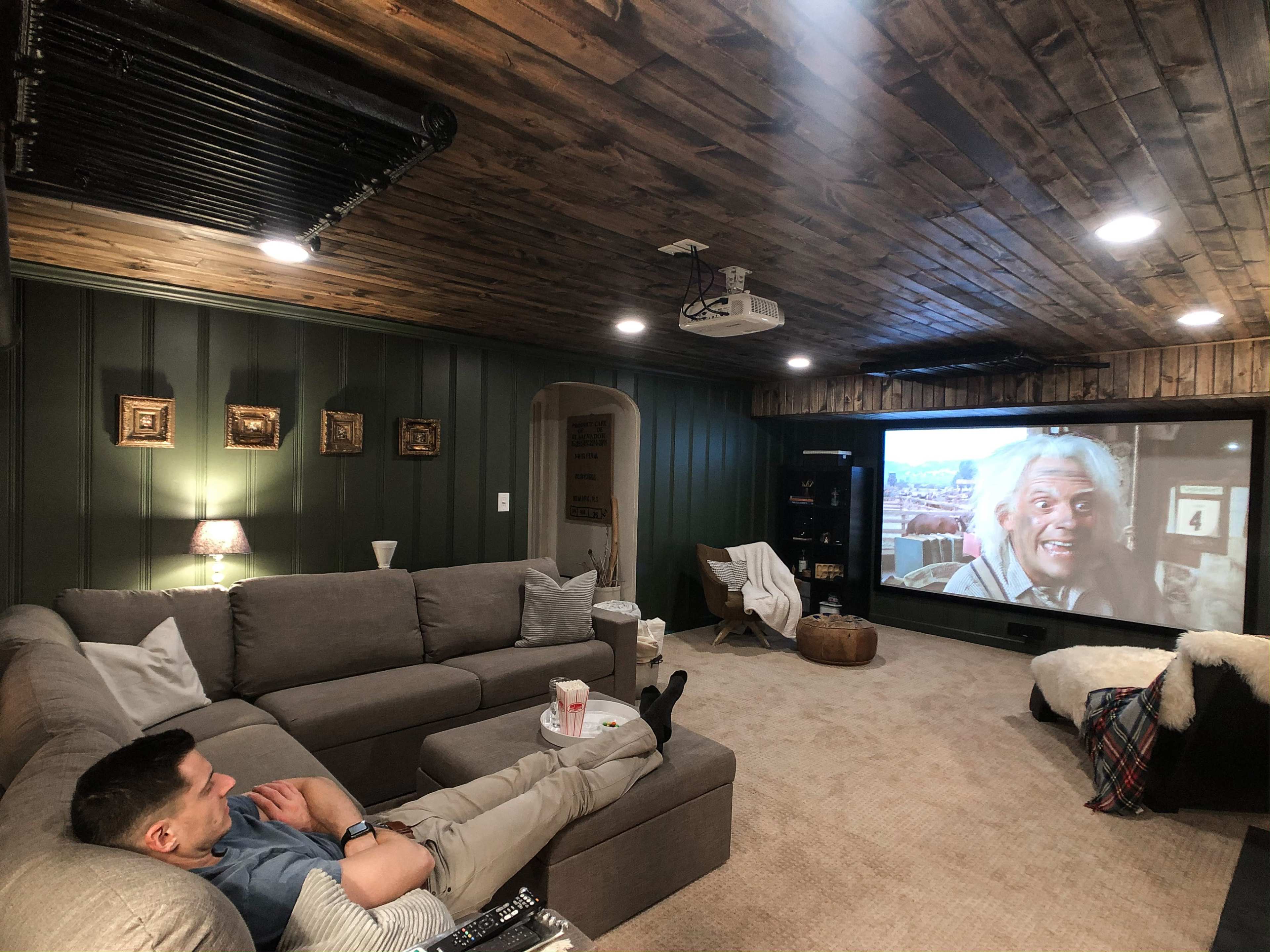 A person reclines on a gray sectional couch in a cozy basement watching a movie on a large projection screen.